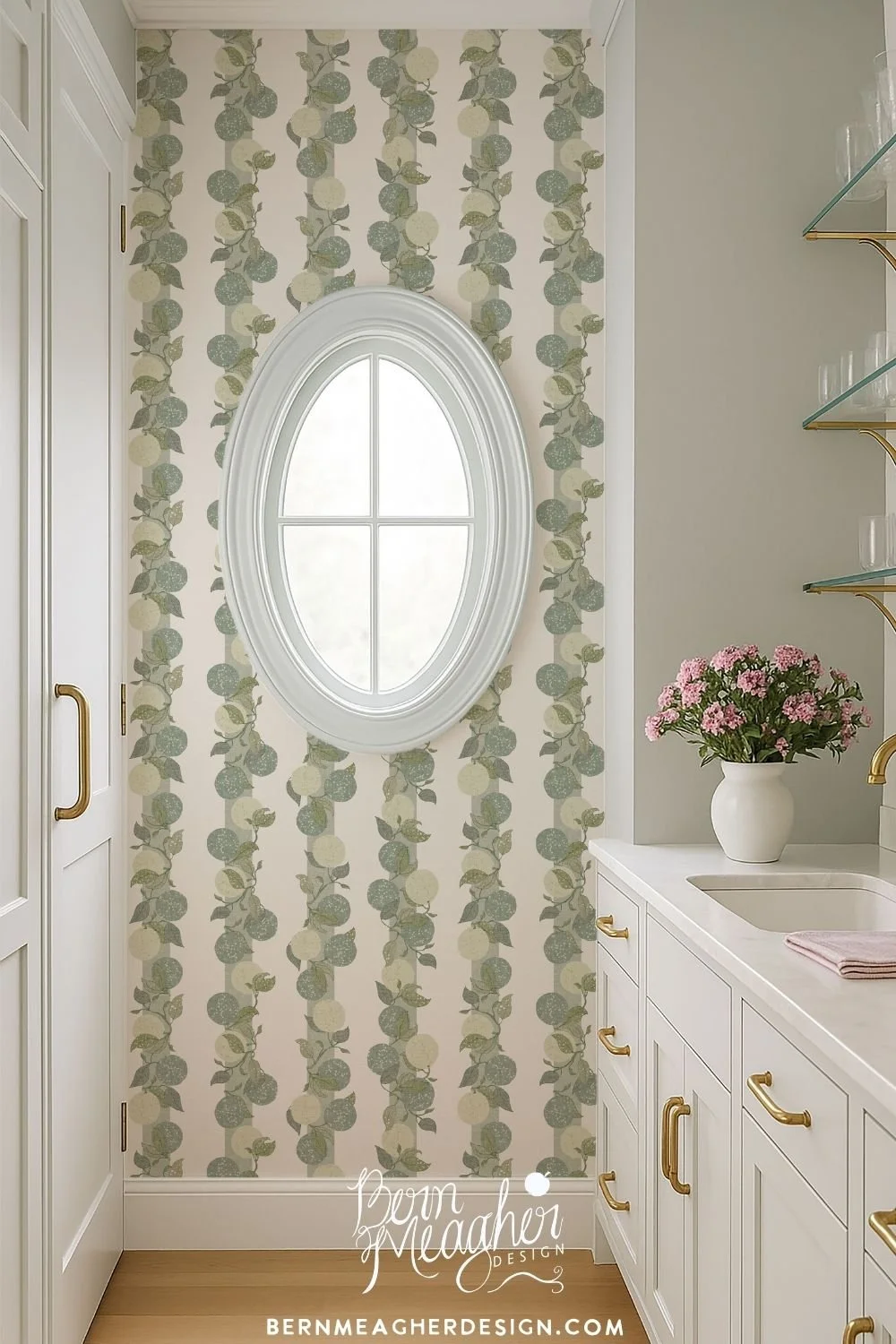 Citrus lime wallpaper in a soft green and cream botanical stripe design with vertical leafy vines. Shown in a bright pantry featuring white cabinetry with brass hardware, a white oval window frame, and a vase of pink flowers on a white countertop.