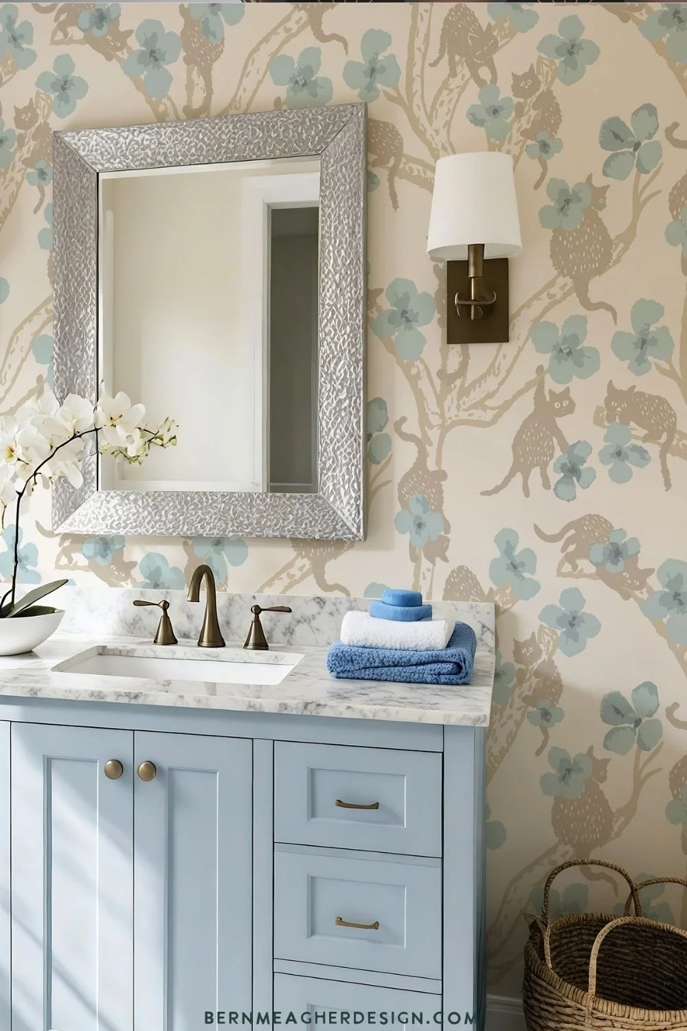 Vinyl bathroom wallpaper with beige branches, playful taupe cats, and blue flowers on a cream background behind a light blue vanity with marble countertop, brass faucet, woven baskets, and a silver framed mirror.