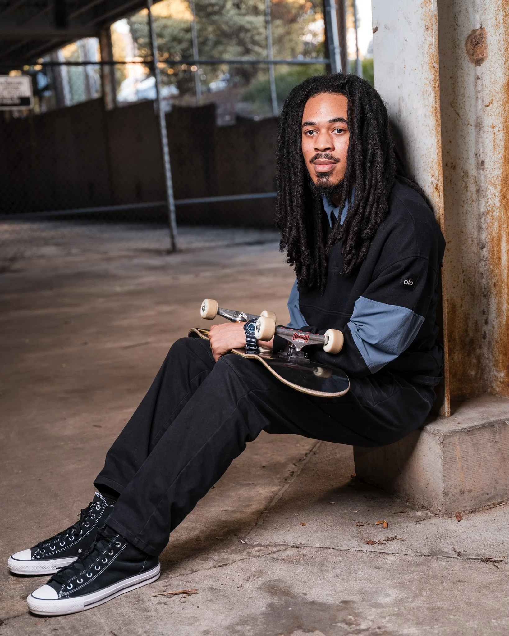 Young man with dreadlocks sitting against a concrete wall in a skate park, holding a skateboard, wearing black high-top sneakers, black pants, a blue shirt, and a black jacket.