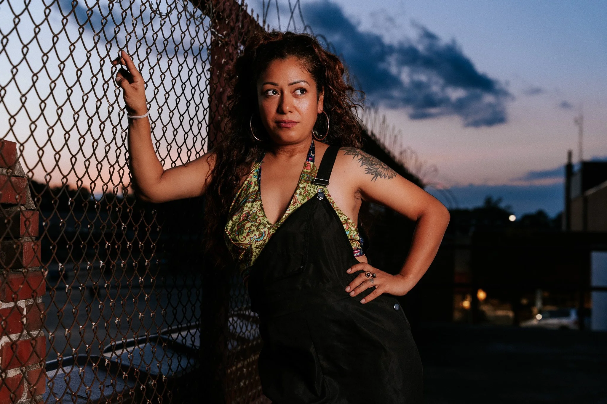 A woman with curly hair and hoop earrings standing next to a chain-link fence during the evening with a cloudy sky in the background.
