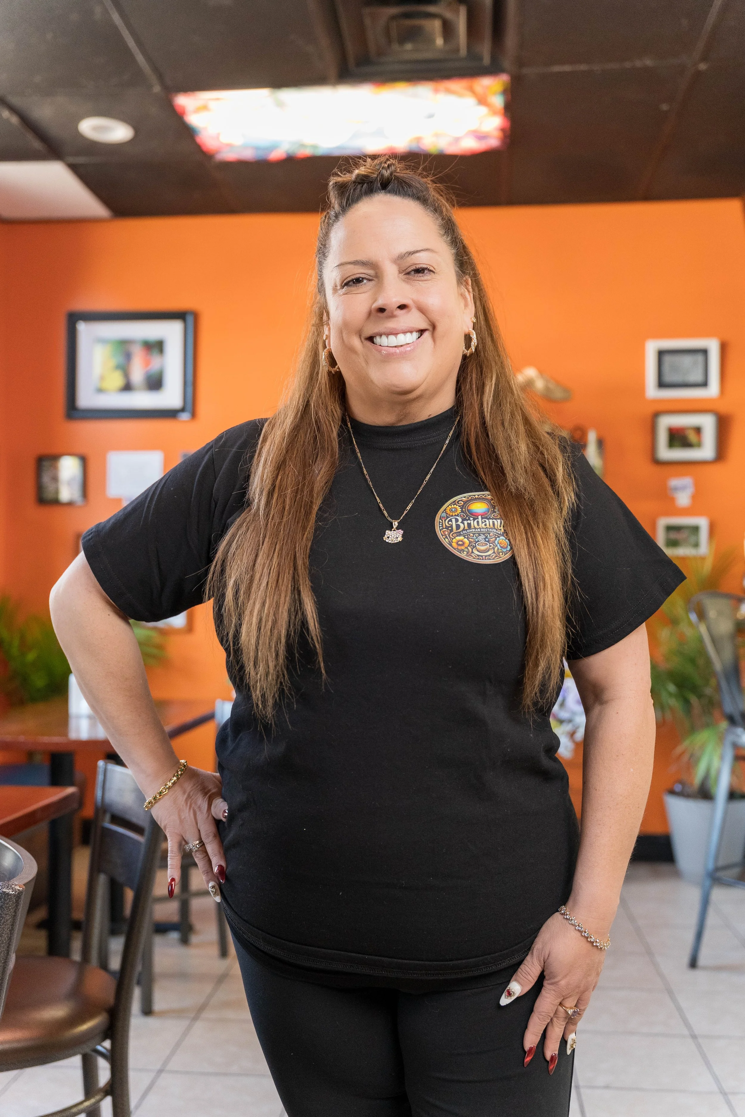 A woman with long brown hair styled in a half-up bun, smiling and posing with her hand on her hip in a restaurant with an orange wall, framed pictures, and tables and chairs in the background.