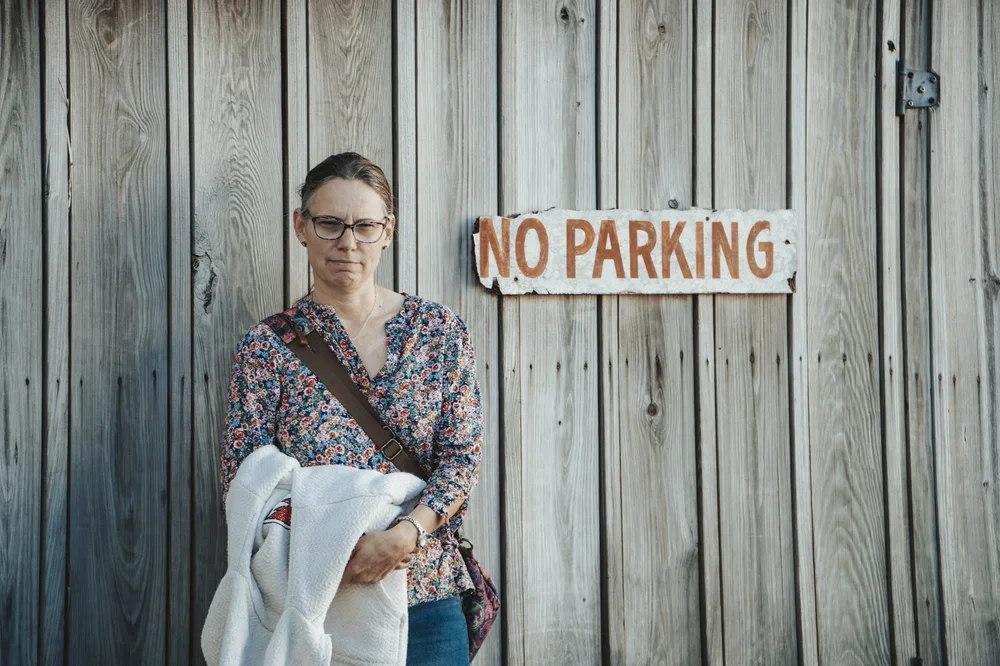 A woman with glasses and a floral shirt standing in front of a wooden fence with a 'No Parking' sign on it, holding a white jacket.