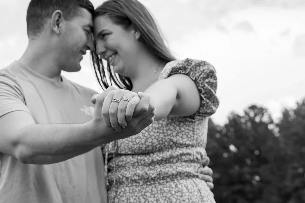 A couple holding hands, with their foreheads touching, smiling at each other outdoors.