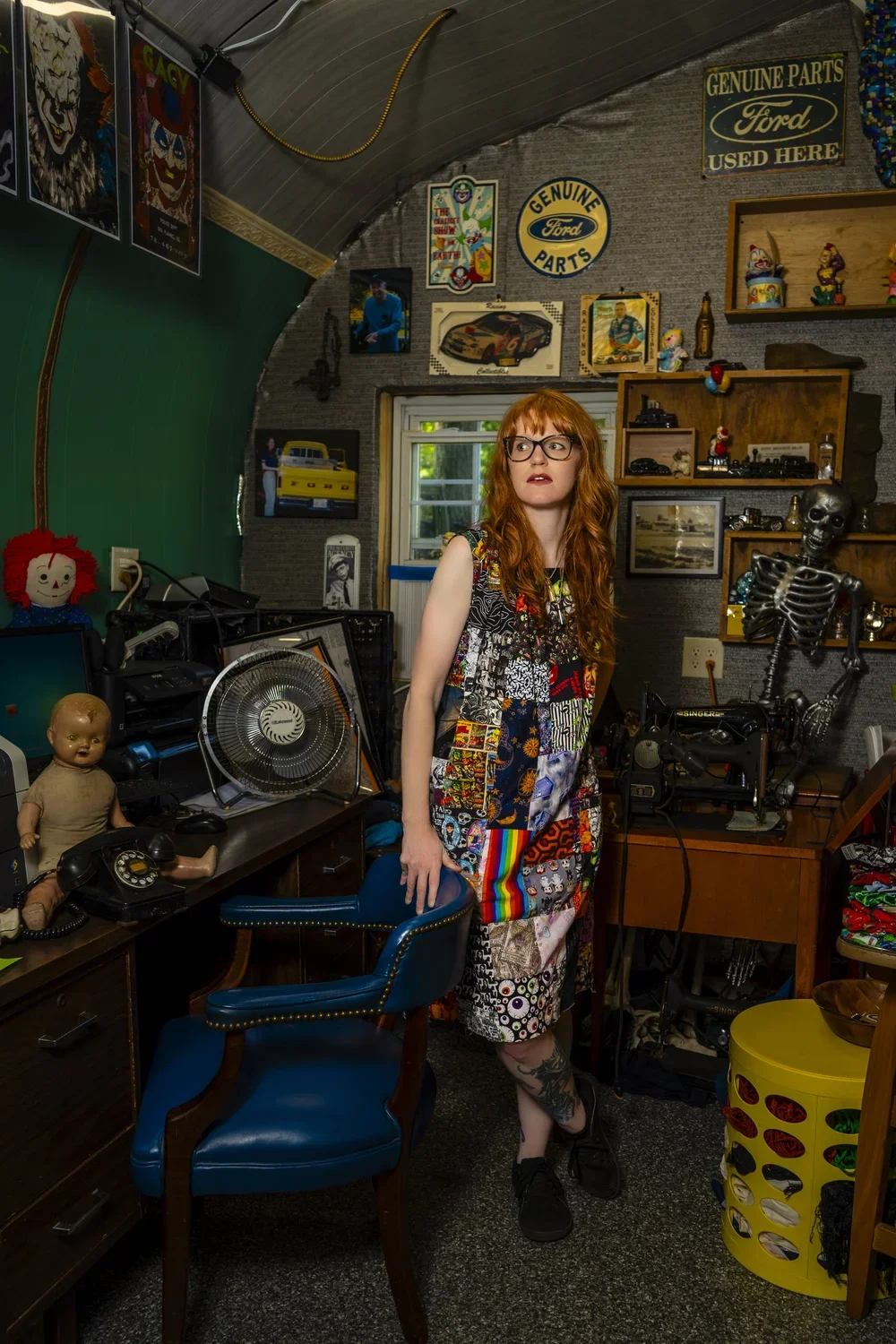 A young woman with red hair and glasses standing in an eclectic room filled with vintage toys, posters, and decorative items, including a skeleton model, a doll, a fan, and various framed pictures and signs on the wall.