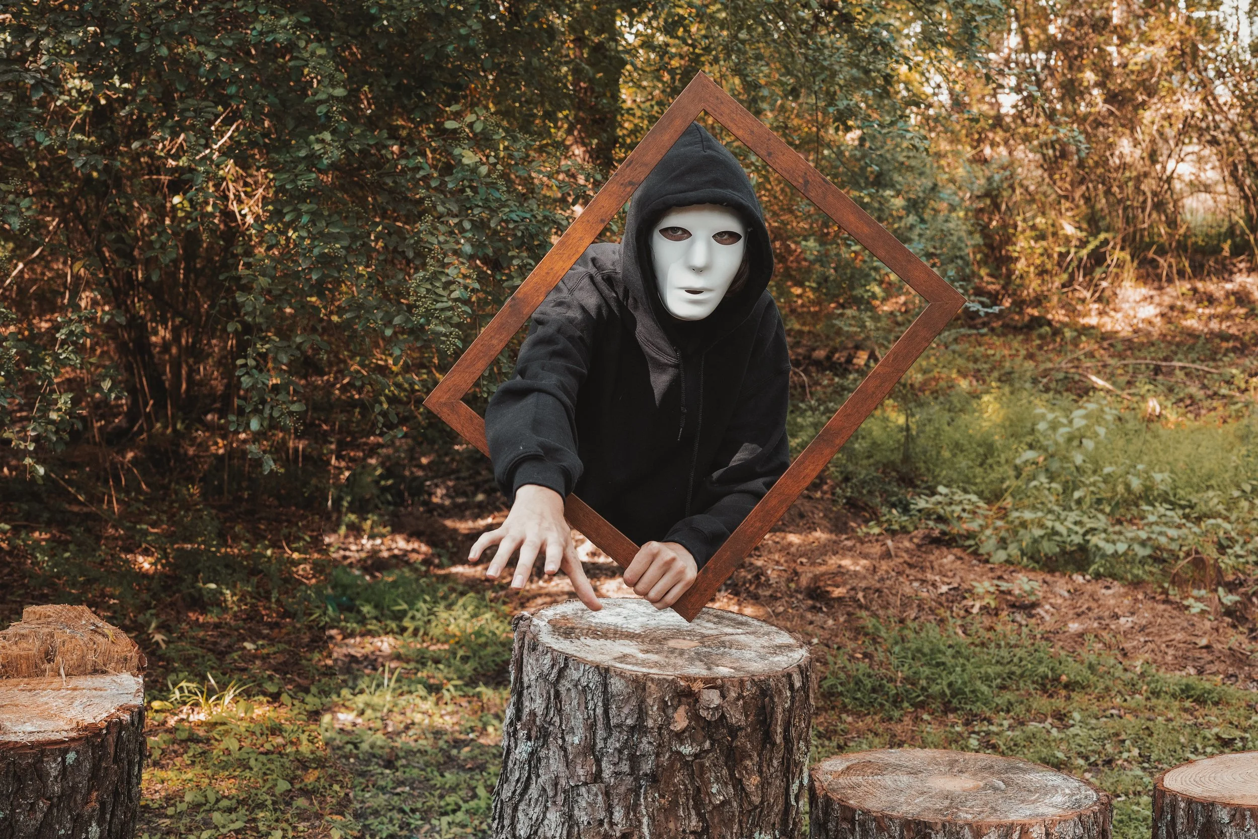 Person wearing a mask and hoodie, reaching out with hand, framed in wooden square, outdoors in a wooded area with tree stumps.