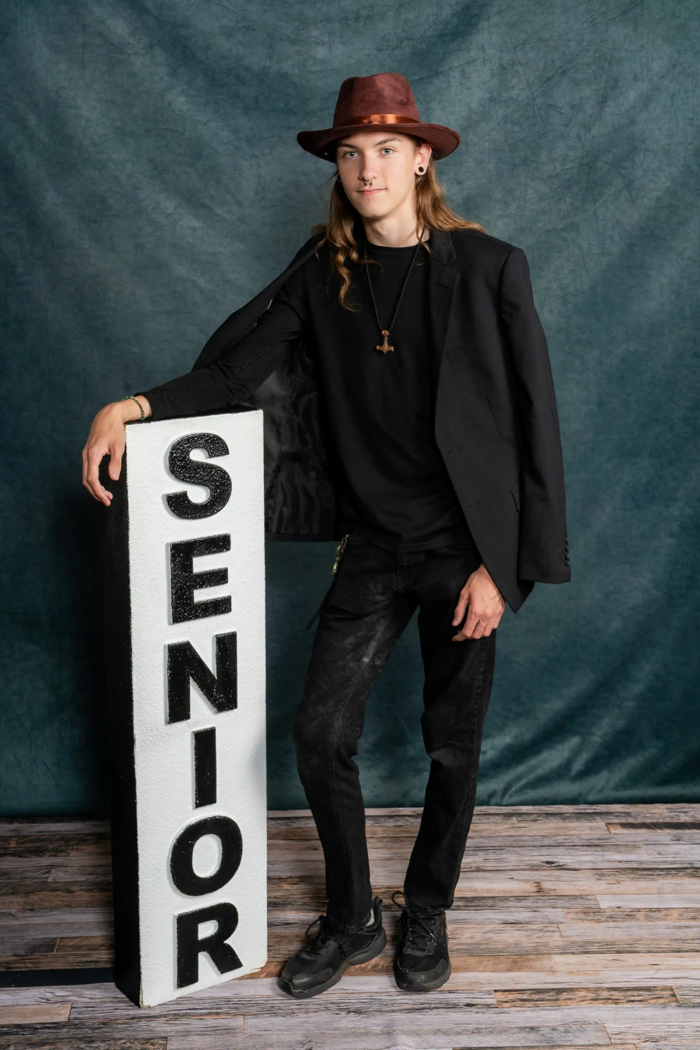 A person with long hair, dressed in a black blazer, black pants, and black shoes, standing next to a tall white sign with the word 'SCHOOL' in black letters, against a blue textured background.