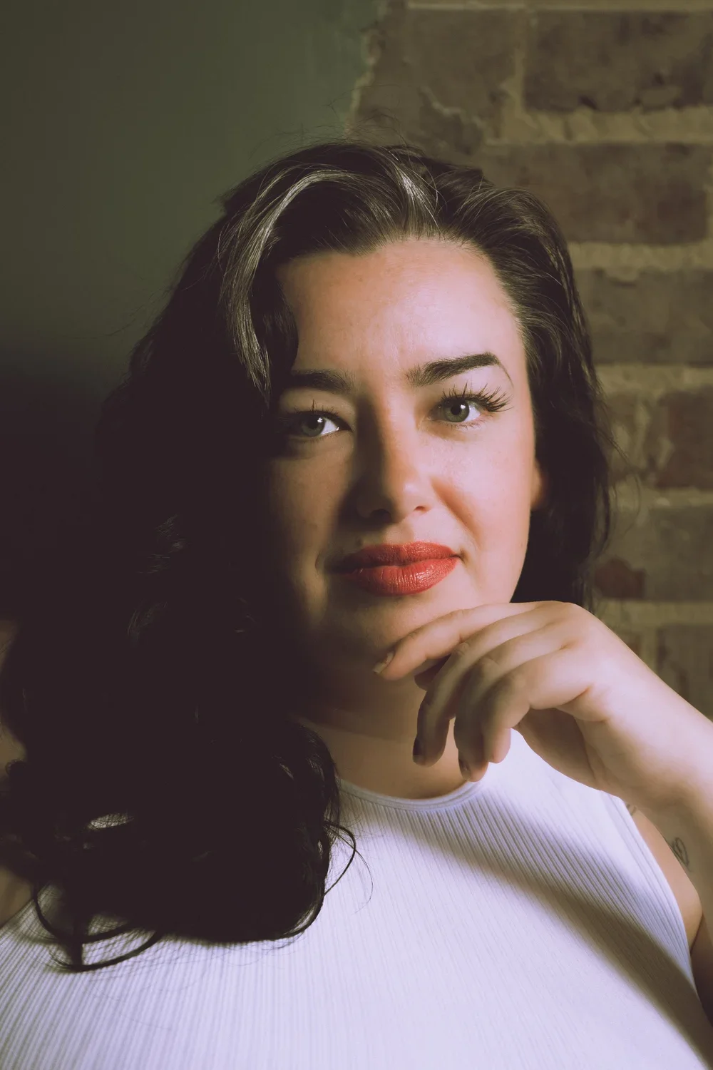 Close-up portrait of a woman with dark wavy hair, red lipstick, and green eyes, resting her chin on her hand, against a brick wall background.