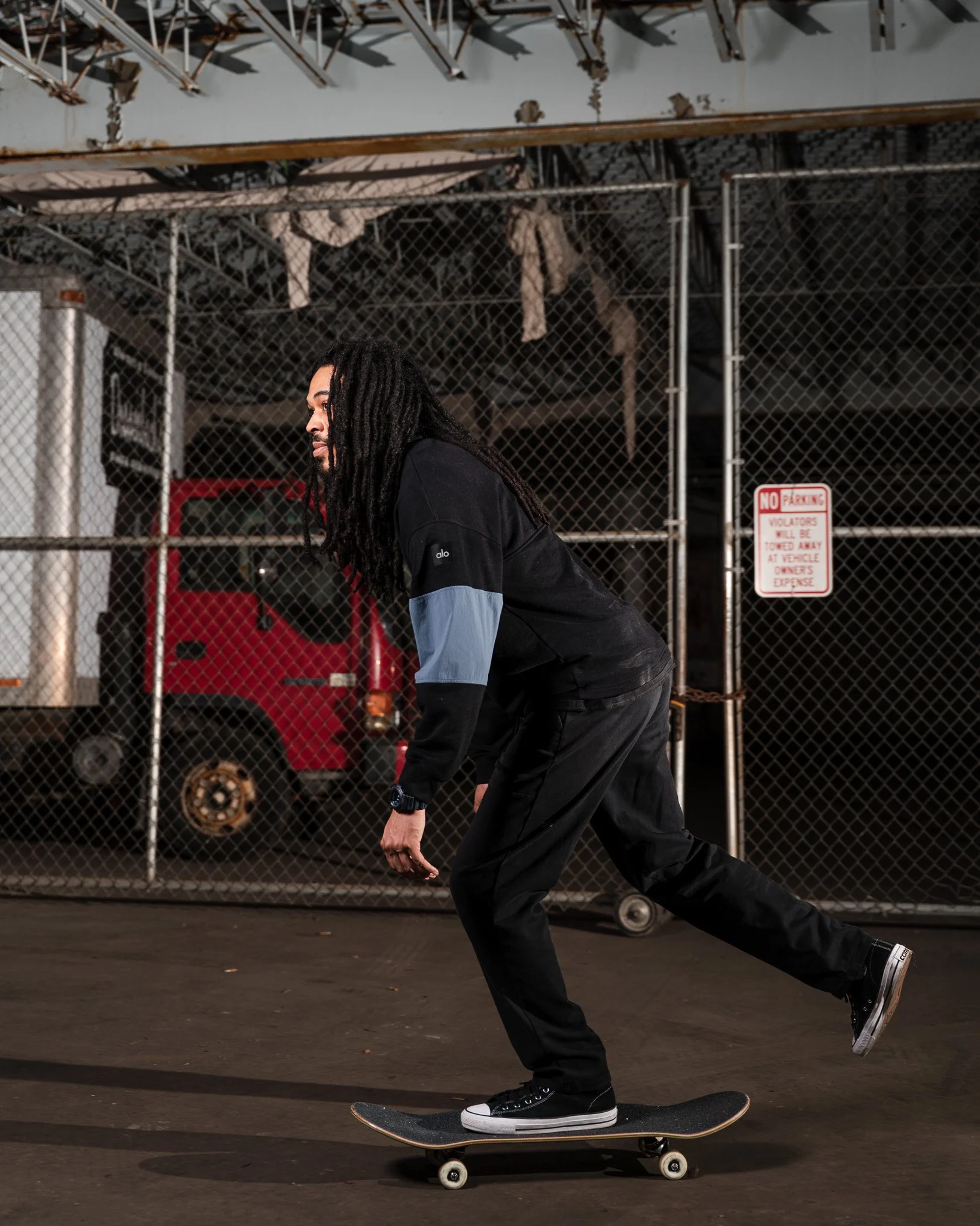 A person with long dreadlocks skateboarding on a flat surface in front of a chain-link fence with a "No Parking" sign. The individual is wearing a black and gray jacket, black pants, and black sneakers, and is captured in Motion.