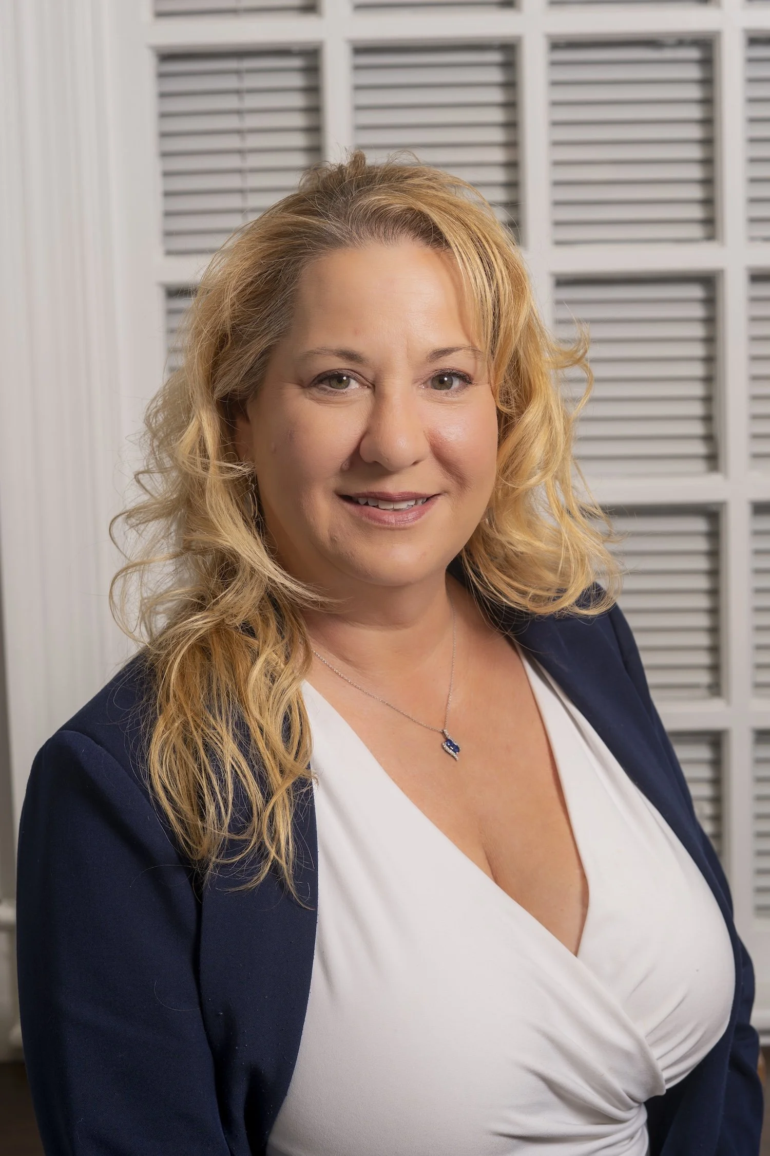 A woman with blonde, wavy hair wearing a white top and navy blazer, smiling at the camera, in front of a window with white plantation shutters.