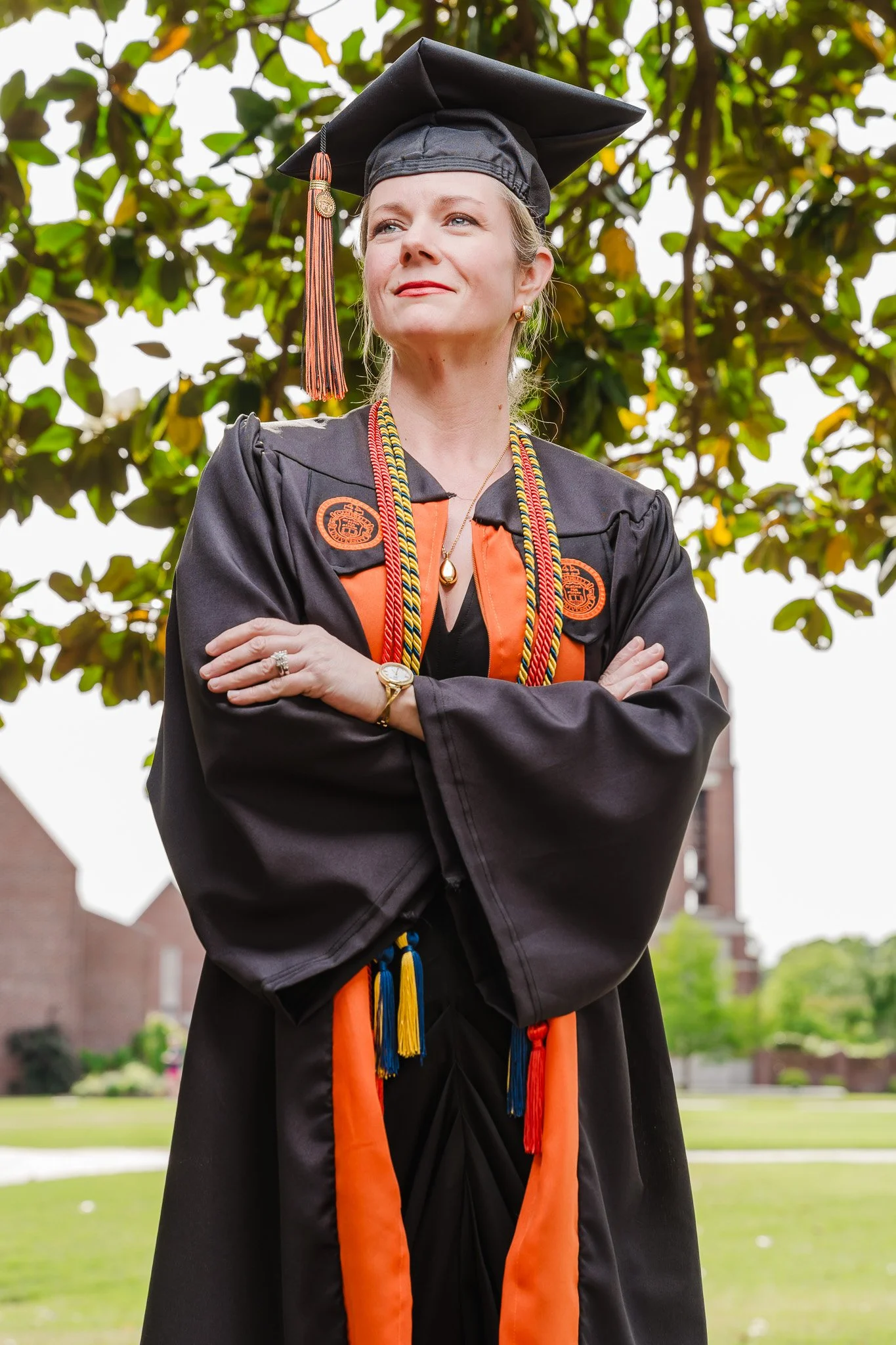 A woman in graduation attire, including a black cap and gown, stands outdoors with her arms crossed, looking confidently into the distance, with green trees and buildings in the background.