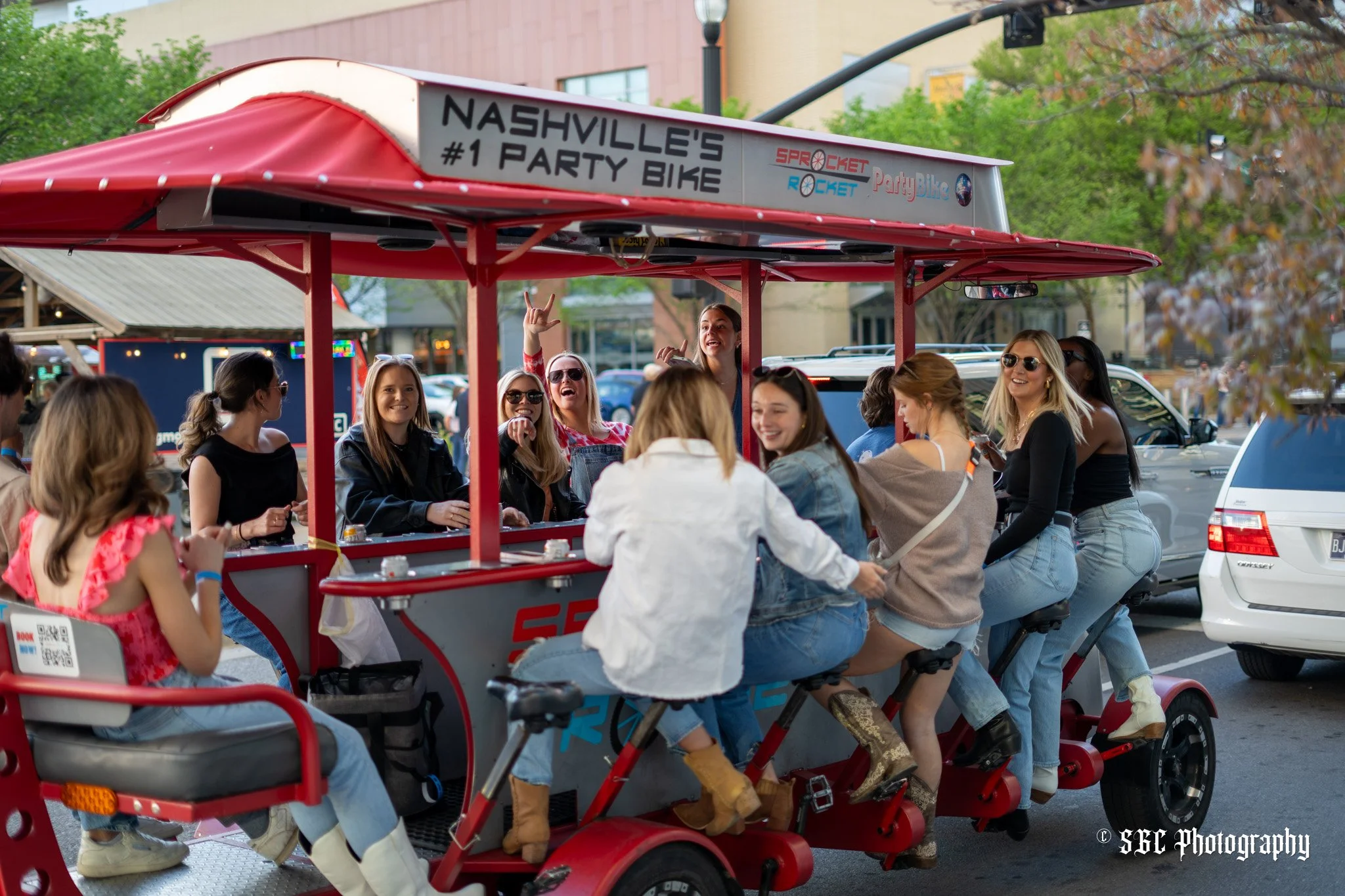 A group of people riding a red three-wheeled bicycle bike with a canopy, with several women smiling and laughing, some wearing sunglasses, parked on a city street with trees and cars in the background.