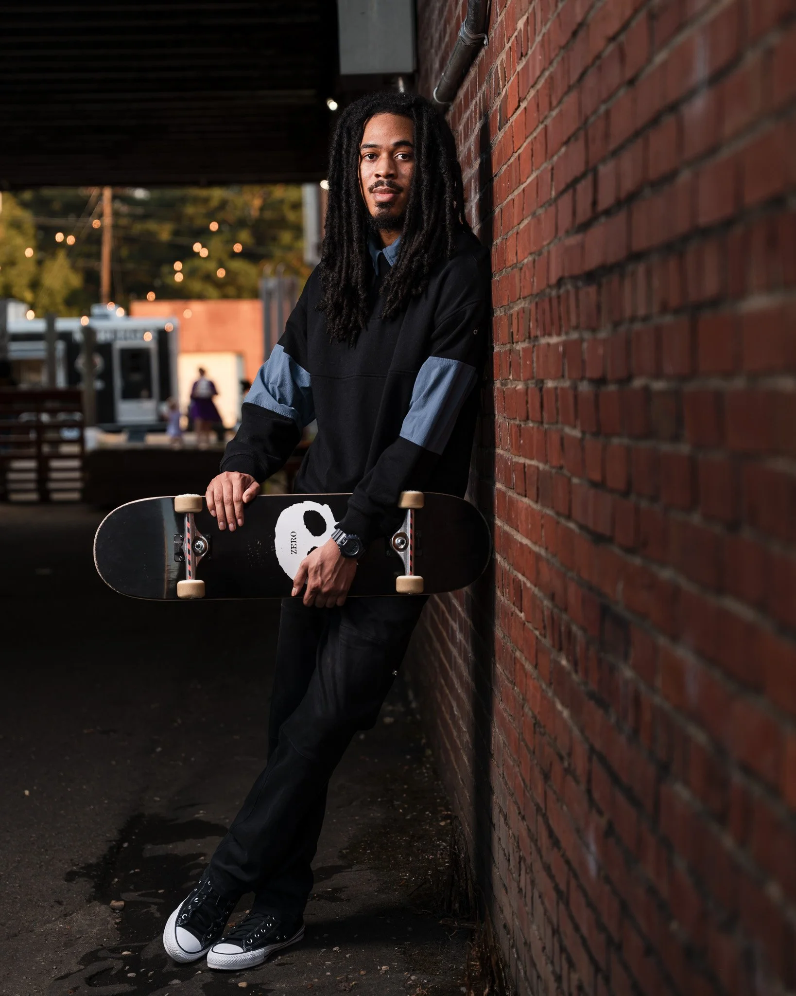 A man with long dreadlocks standing next to a brick wall outside during the evening, holding a skateboard and looking at the camera.