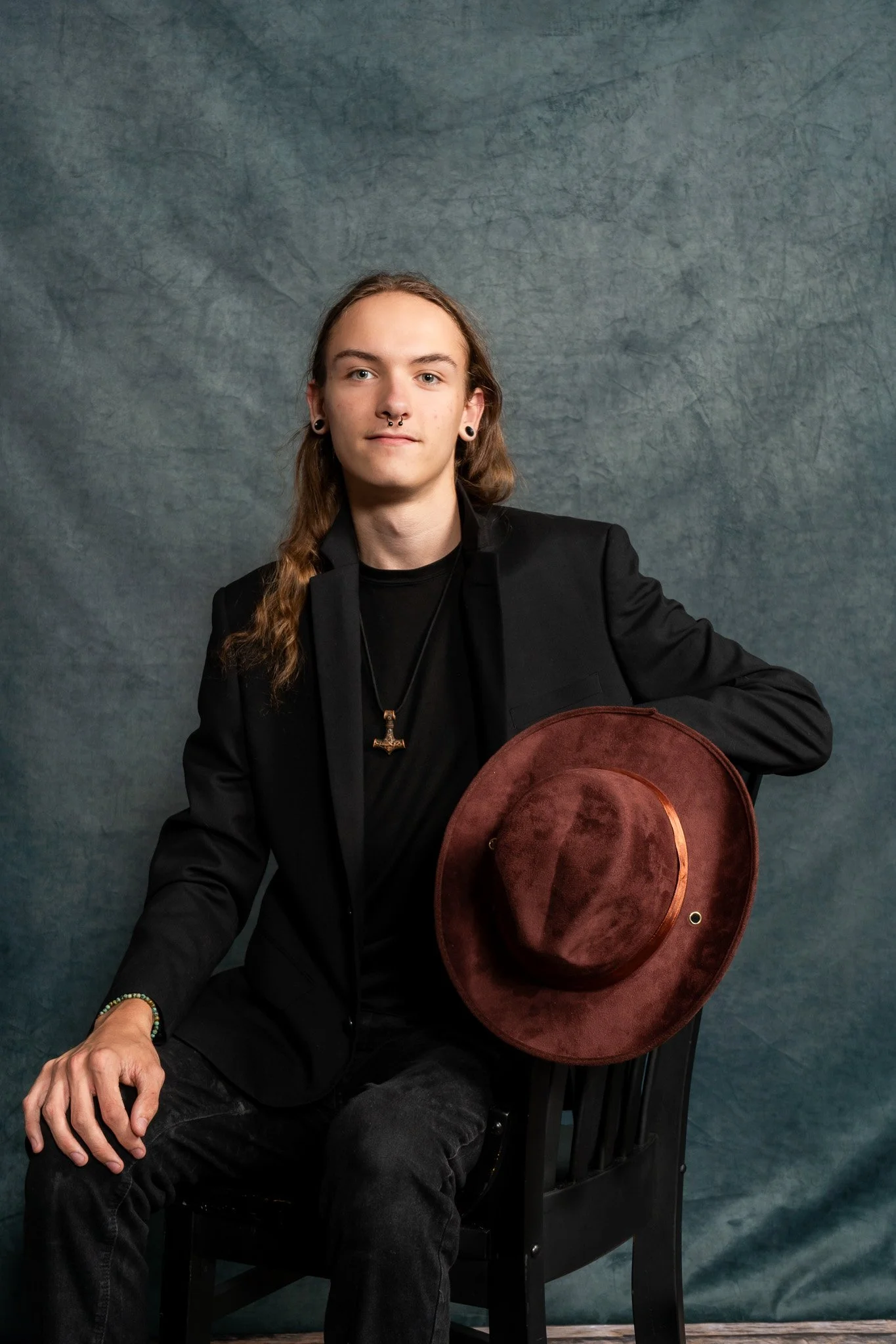 A young person with long brown hair, earrings, and a septum piercing, wearing a black blazer and shirt, sitting on a black chair, holding a wide-brimmed brown hat, against a textured blue-gray background.