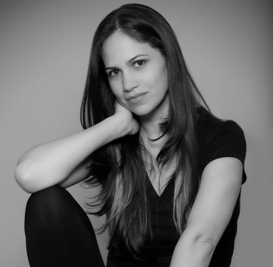 Black and white portrait of a woman with long hair, wearing a black top, sitting with one hand resting on her neck.
