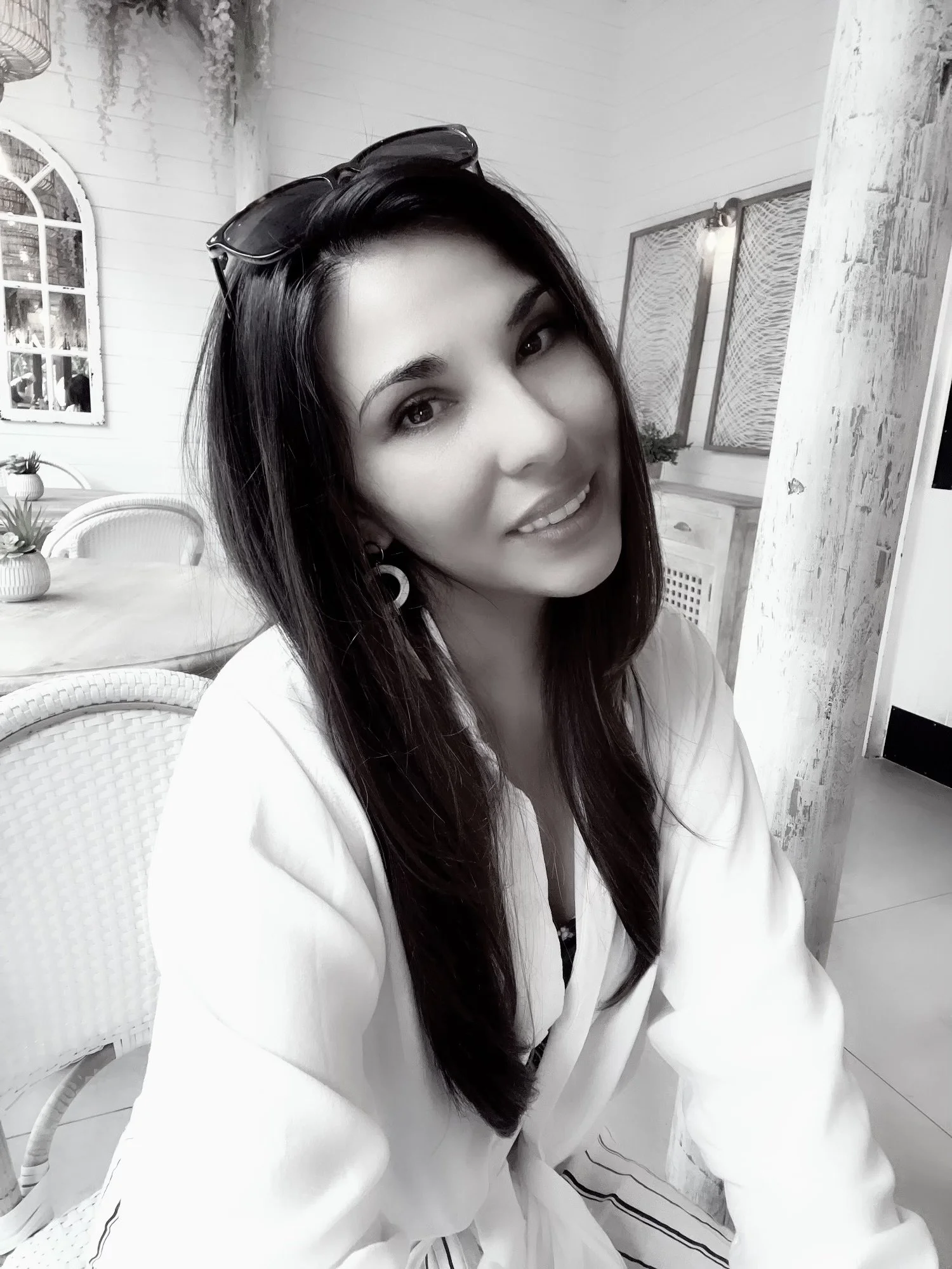 Black and white portrait of a smiling woman with long hair, wearing a white top. The setting is a cozy room with wicker chairs and decorative elements.