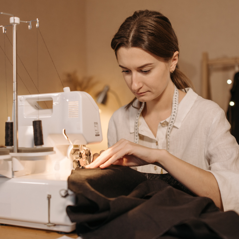 Person sewing fabric using a sewing machine, wearing a measuring tape around the neck, focused on stitching.