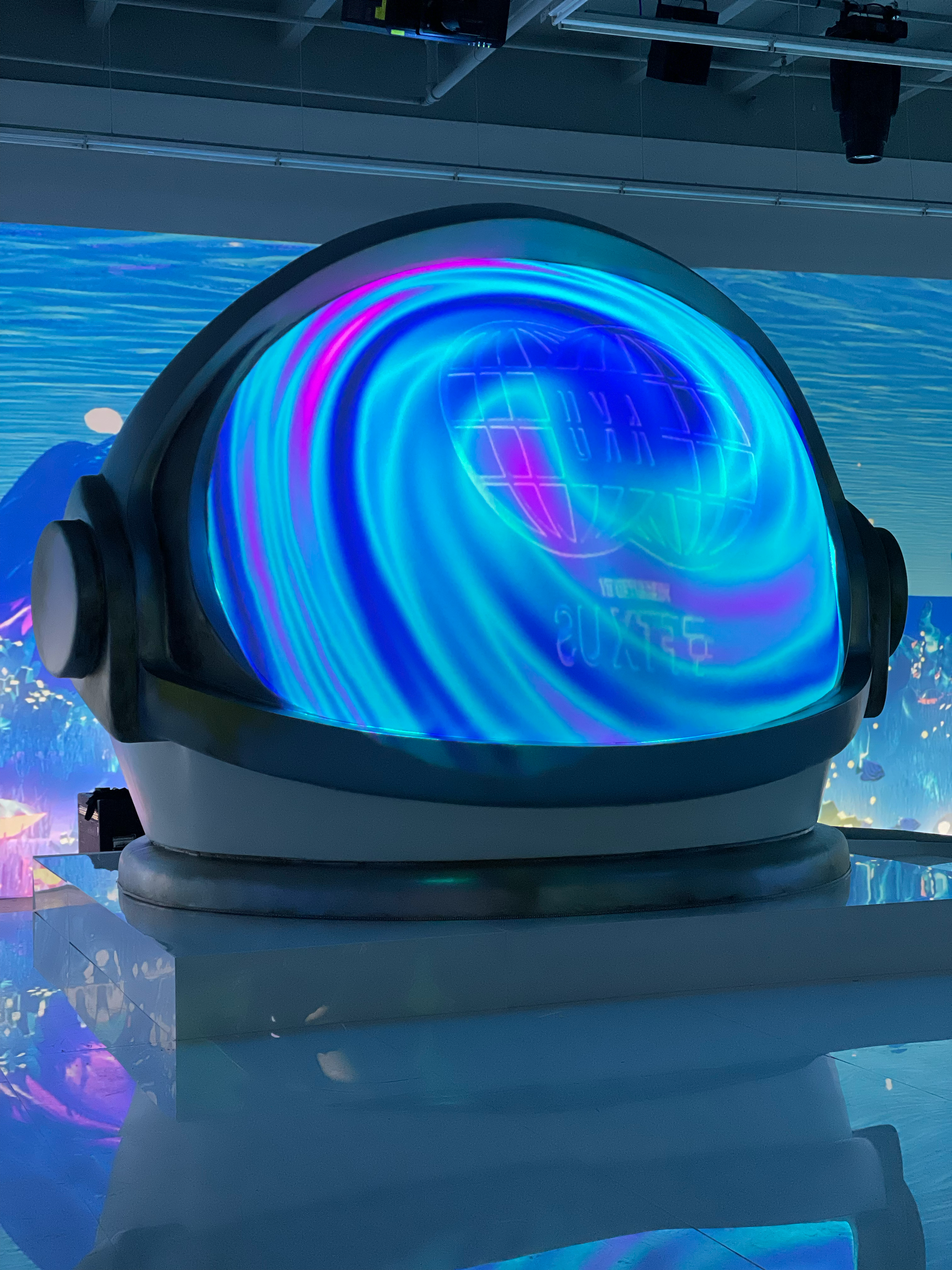 A large digital device with a circular screen displaying colorful, swirling lights and patterns, set in an indoor environment with a background of marine-themed murals.