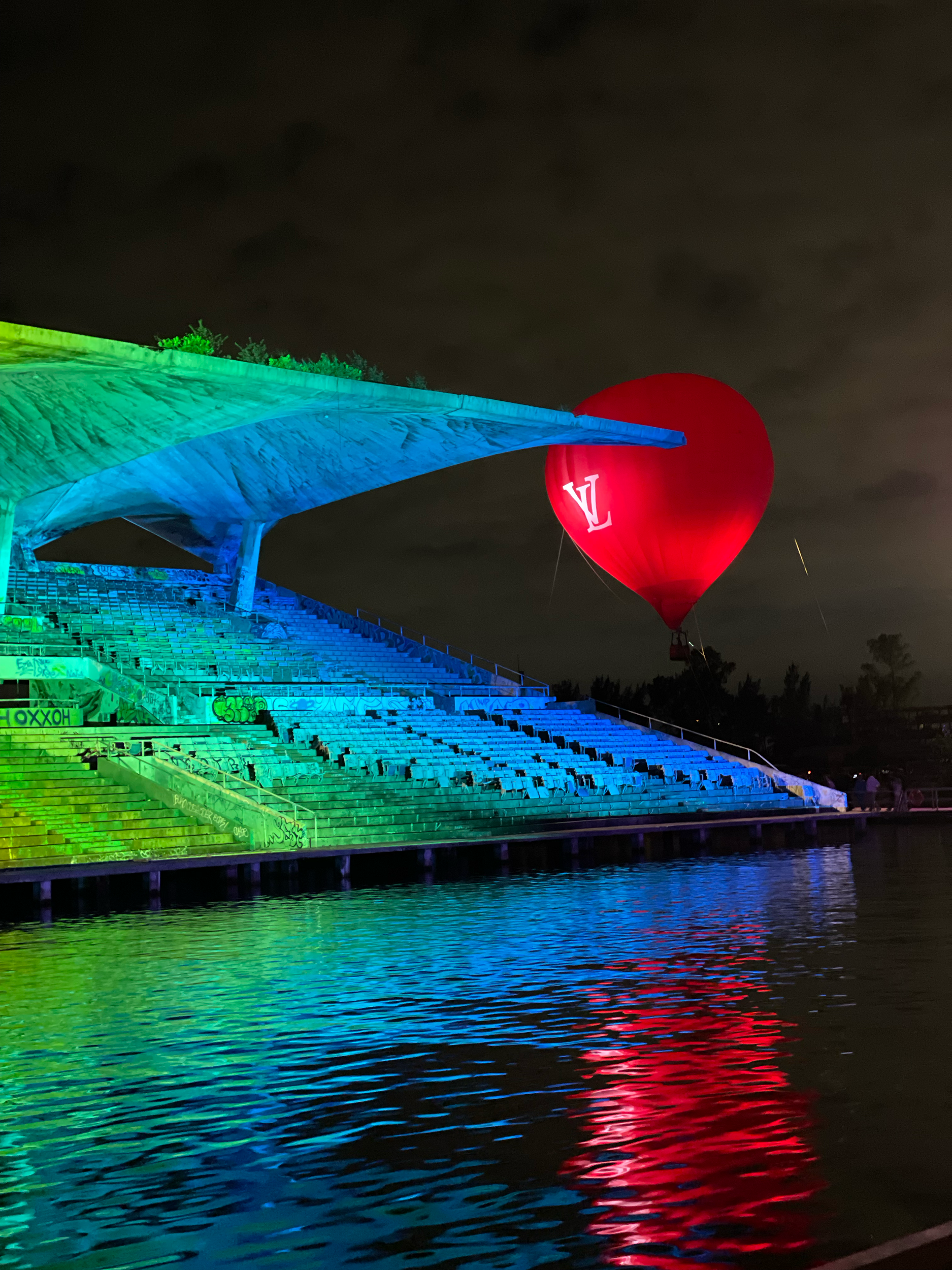 Nighttime view of a waterway with a concrete amphitheater or stadium steps illuminated with colorful lights in green, blue, and red. A large red hot air balloon with the Louis Vuitton logo floats in the sky above, against a dark cloudy sky.