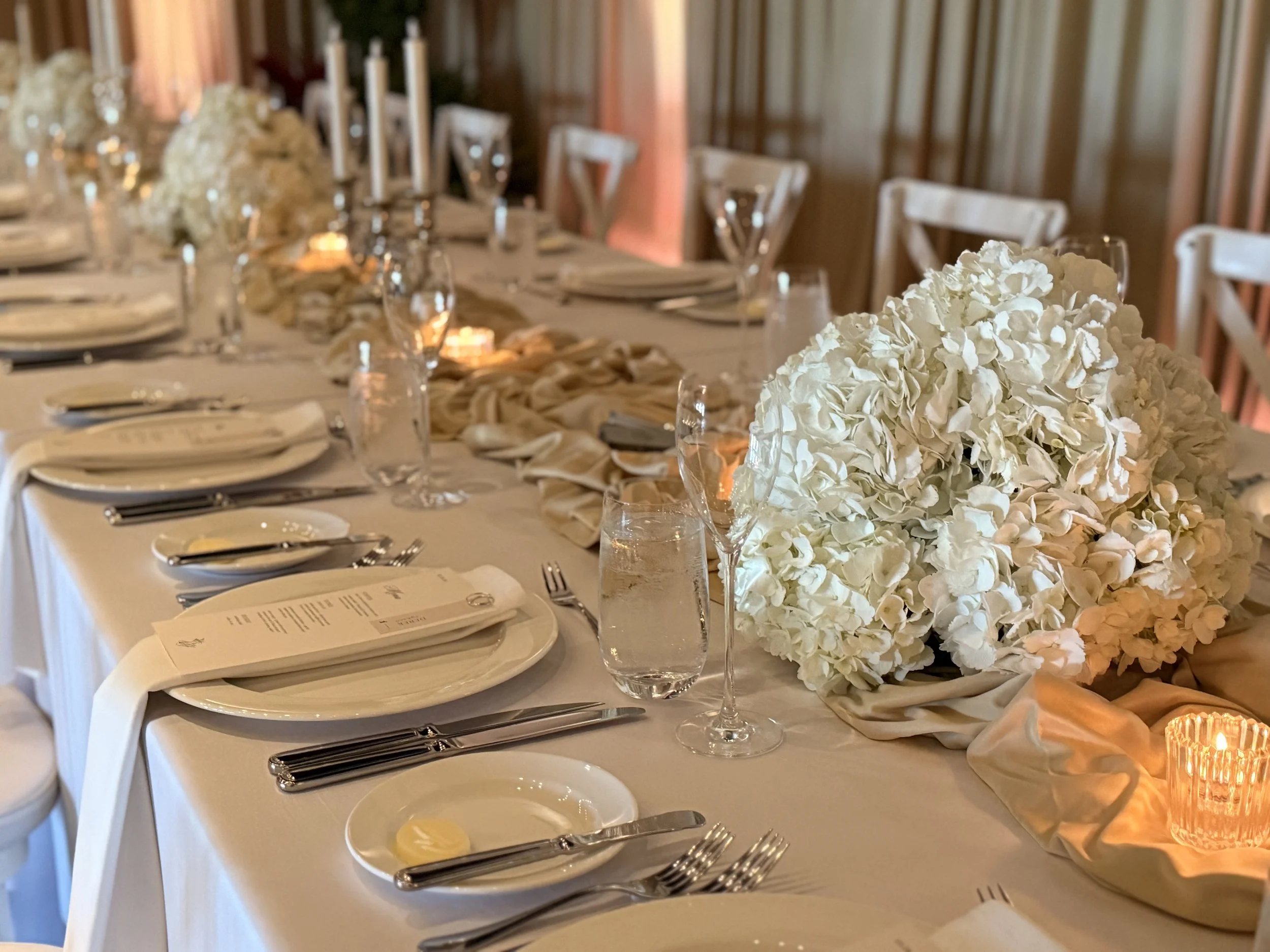 A formal dining table set for a wedding or special event, with white floral centerpieces, glasses, silverware, and candles, in an elegantly decorated room.