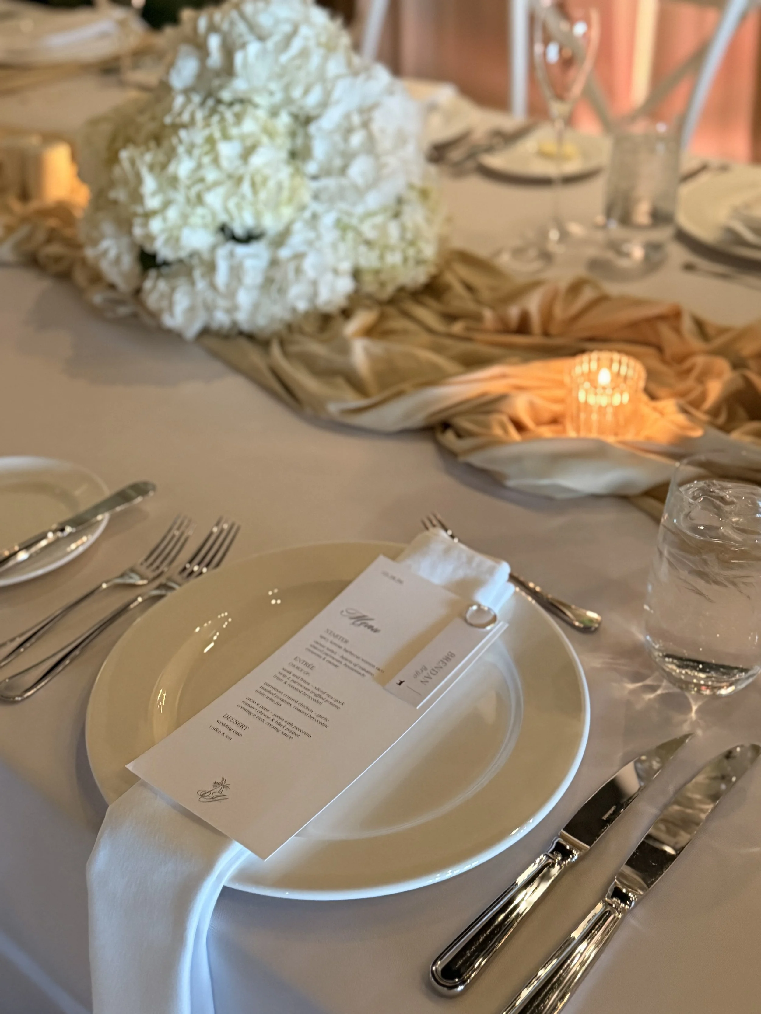 Table setting at a formal event with white tablecloth, plate, utensils, glass of water, and a menu. In the background, a floral centerpiece with white flowers and a small candle.