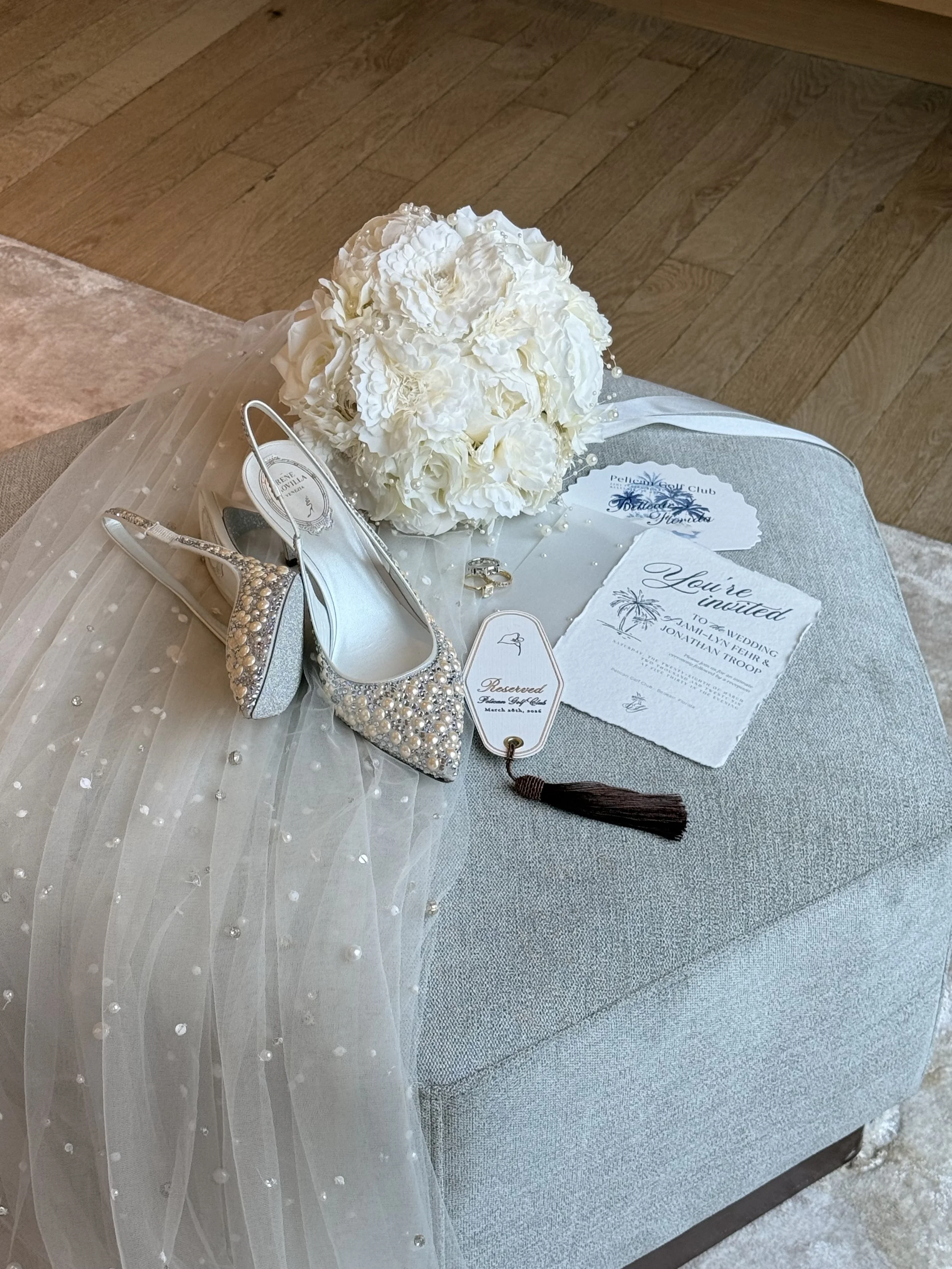A bouquet of white flowers with pearl accents, a pair of silver, rhinestone-embellished shoes with a T-strap design, wedding invitation cards, a ring, and a reserved sign on a gray table cloth, with a wooden floor in the background.