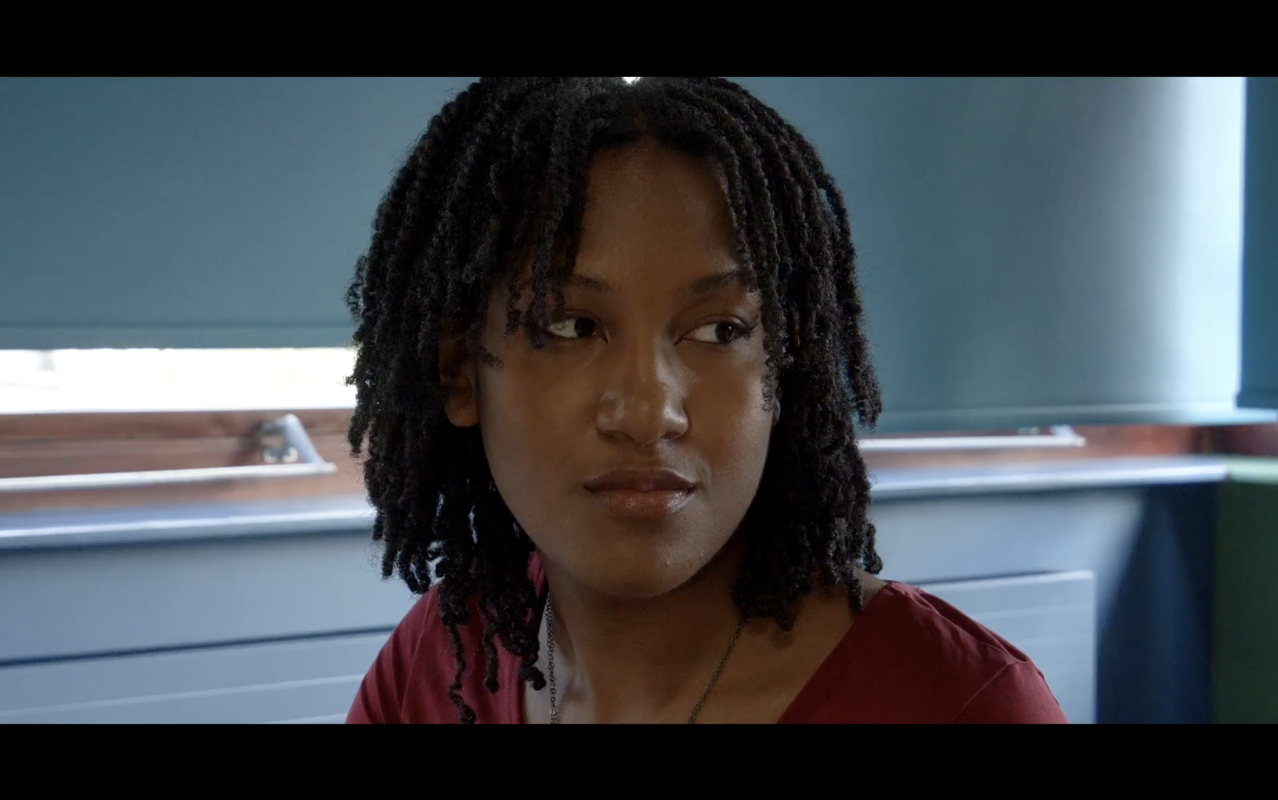 Screenshot from Clara Holland's 'Renascence'. Close-up of a woman with dark natural hair, wearing a maroon shirt, looking to the side with a neutral expression, in a room with a window and blinds in the background.