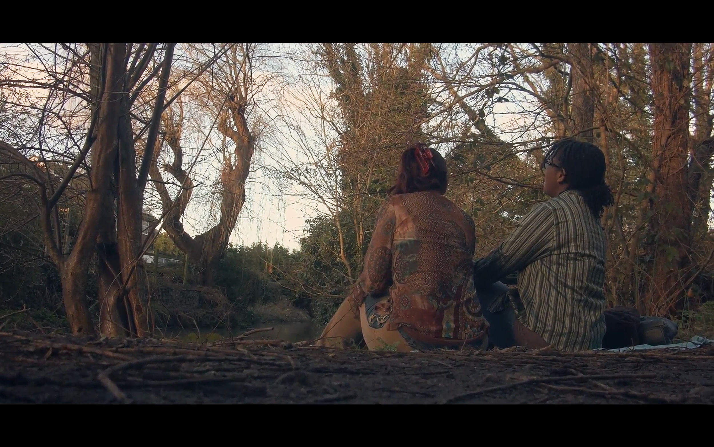 Screenshot from Clara Holland's 'Renascence'. Two people sitting by a small river in a wooded area during sunset, facing away from the camera.