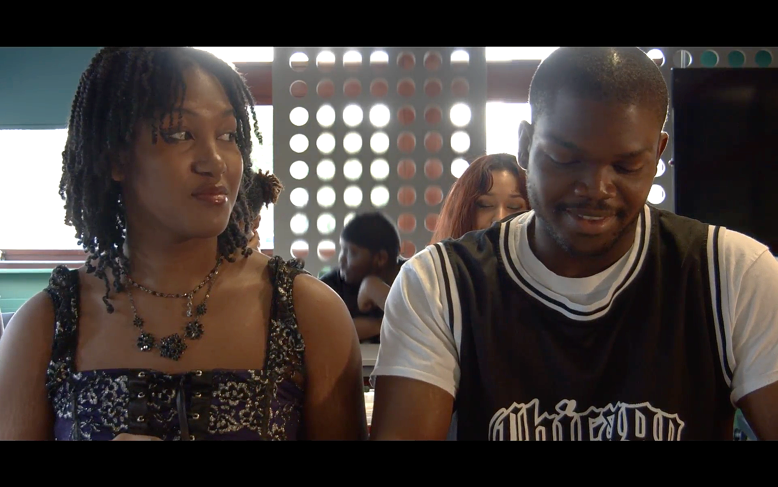 Screenshot from Clara Holland's 'Renascence'. A woman with braids wearing a black and purple dress and jewelry sitting next to a man with short hair and a black and white t-shirt, in a classroom.