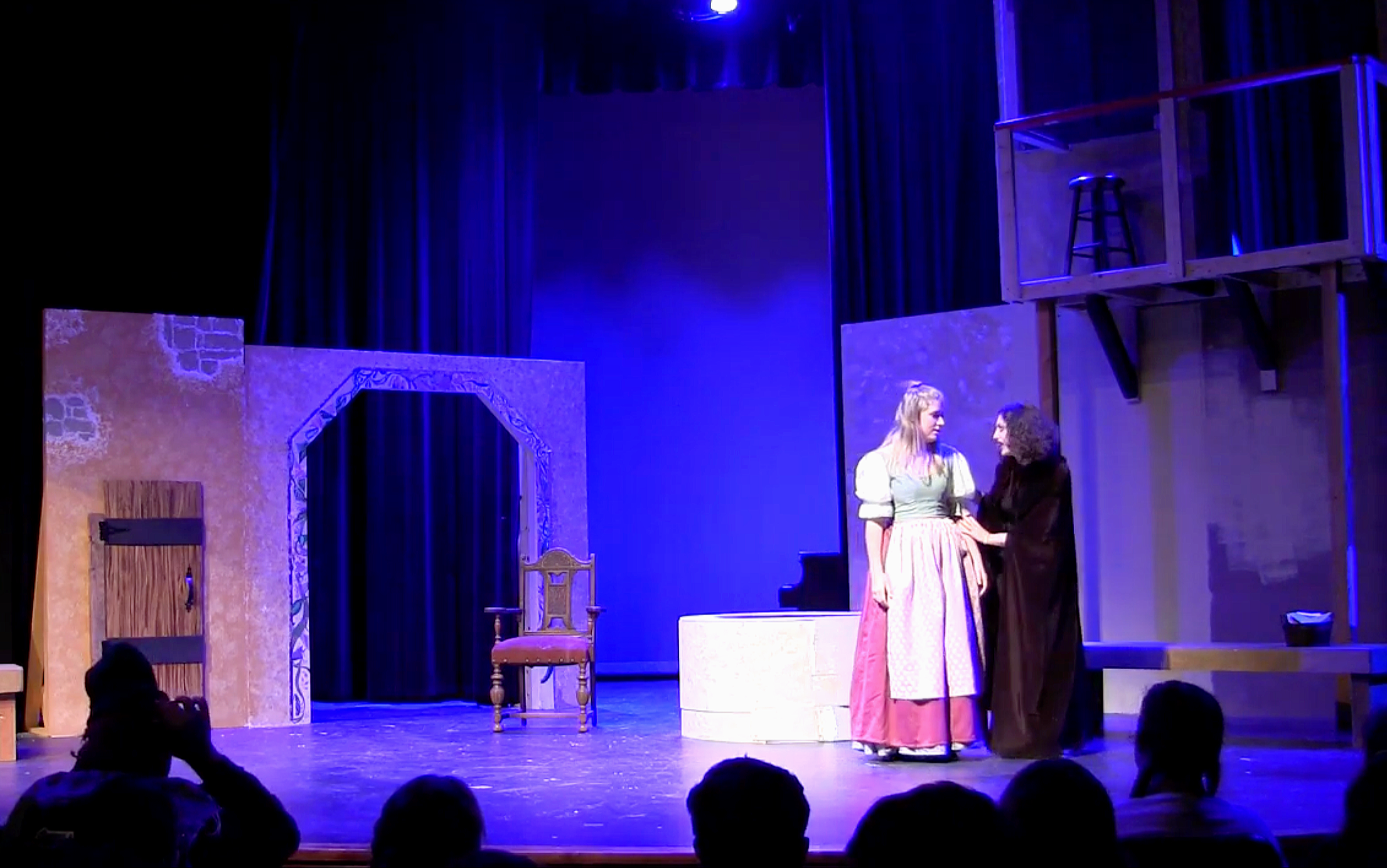 Tableau from Clara Holland's 'Glass Slipper'. The Evil Fairy Godmother confronts Cinderella, with set pieces including a chair, a doorway with a painted stone wall, and a loft with a stool, under blue stage lighting.