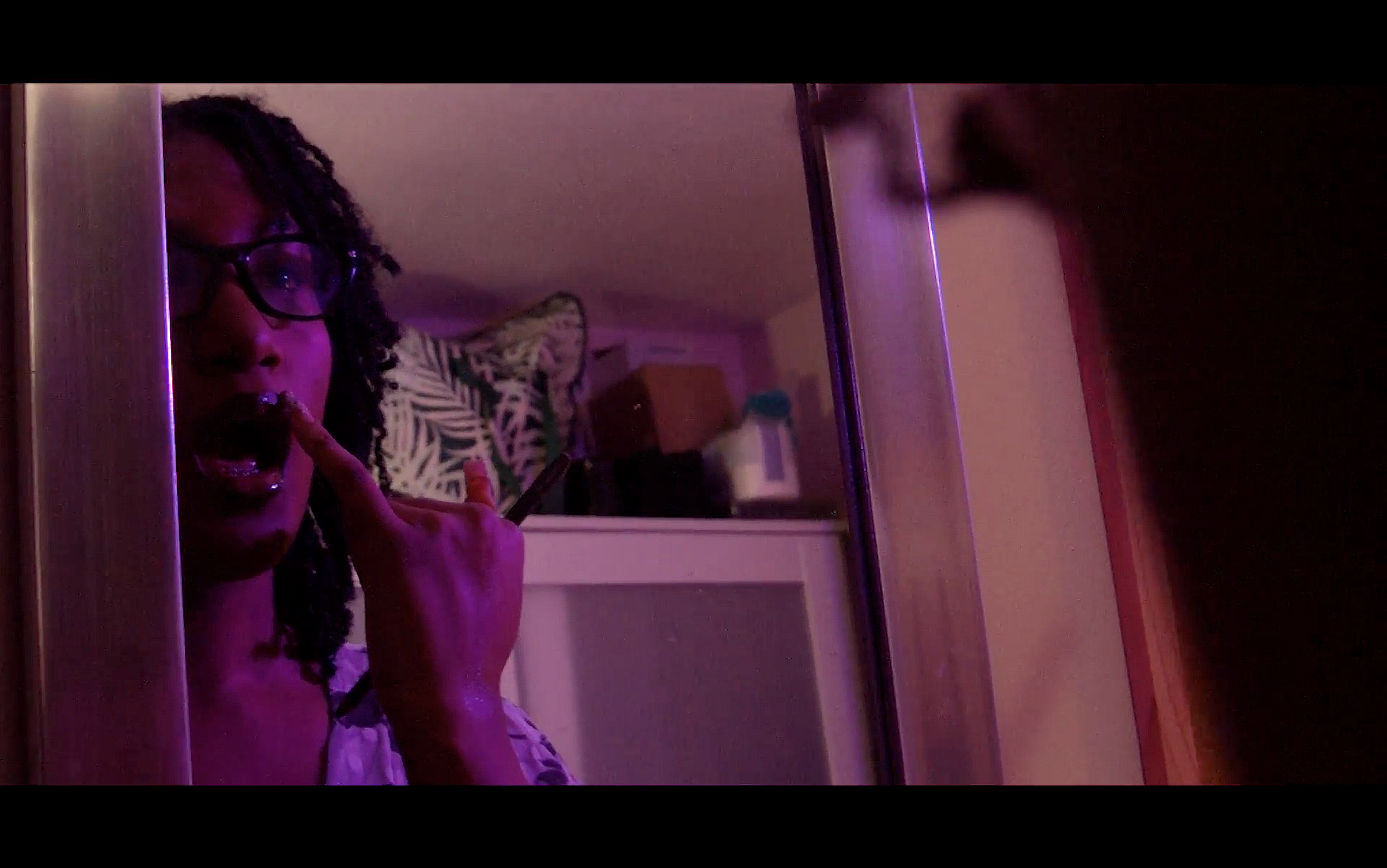 Screenshot from Clara Holland's 'Renascence'. A woman with glasses and braids taking a selfie in a mirror, making a rock sign with her finger, in a dimly lit room with purple lighting.