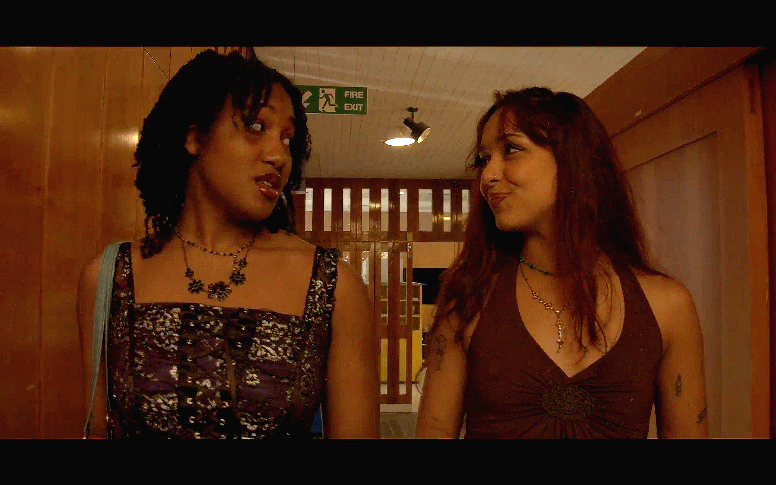 Screenshot from Clara Holland's 'Renascence'. Two women with curly hair walking indoors, one with dark skin and the other with lighter skin, both wearing sleeveless dresses and necklaces, engaging in conversation.