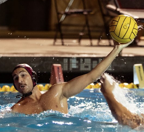 A water polo player in a swimming pool, wearing a cap, reaching out to throw a yellow water polo ball.