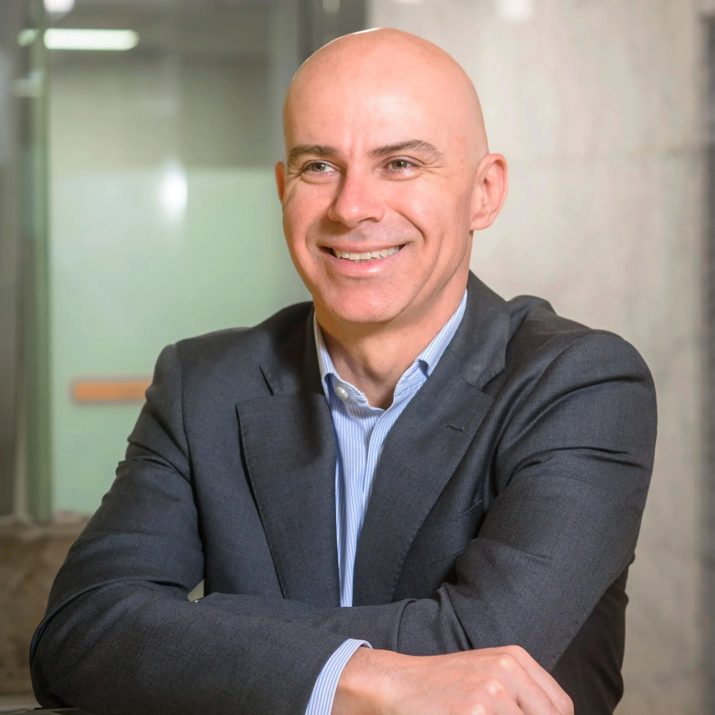 A smiling bald man in a dark suit with arms crossed, sitting at a desk in a professional office environment.
