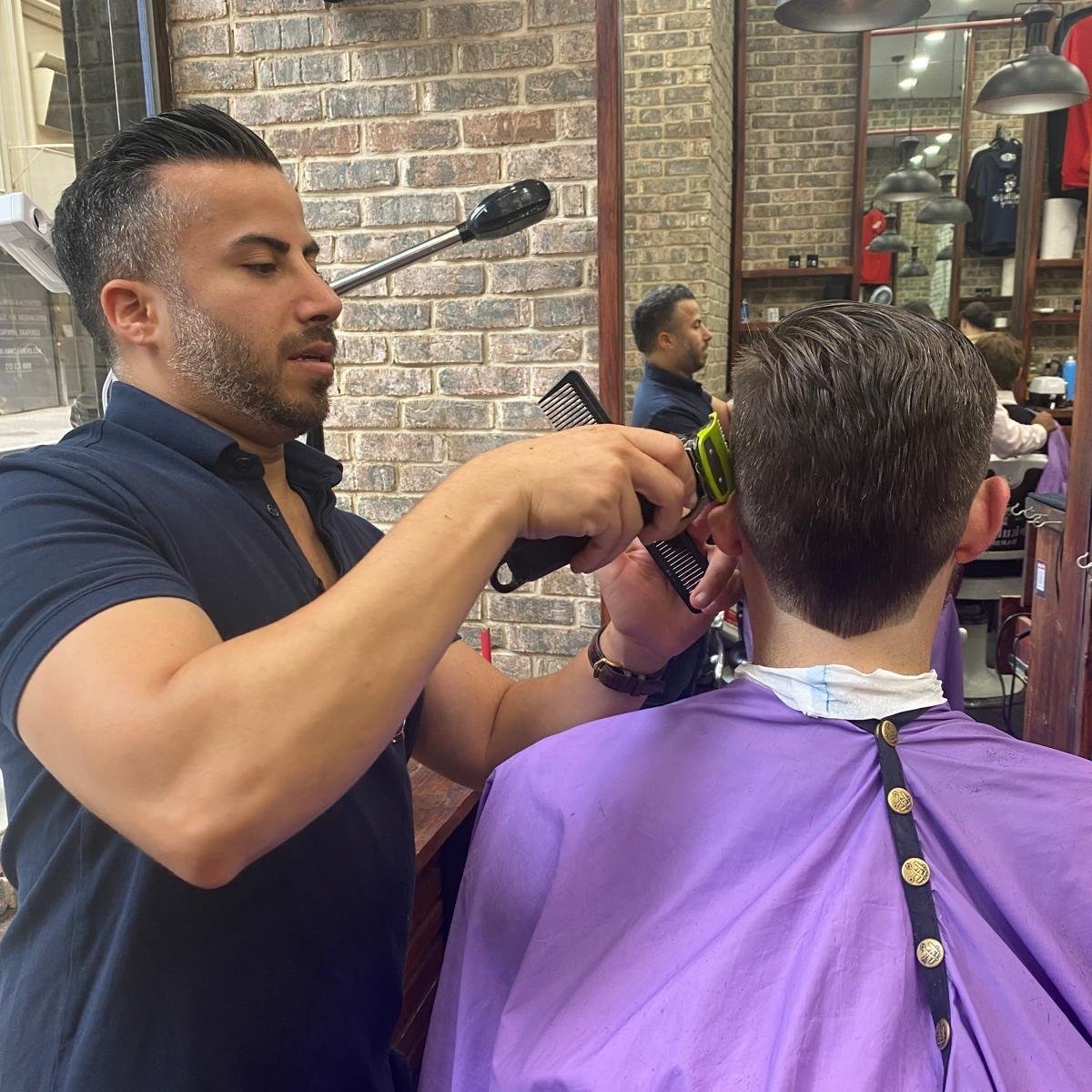 A barber is cutting a man's hair with scissors and a comb in a salon, wearing a purple cape, with mirrors and other customers in the background.
