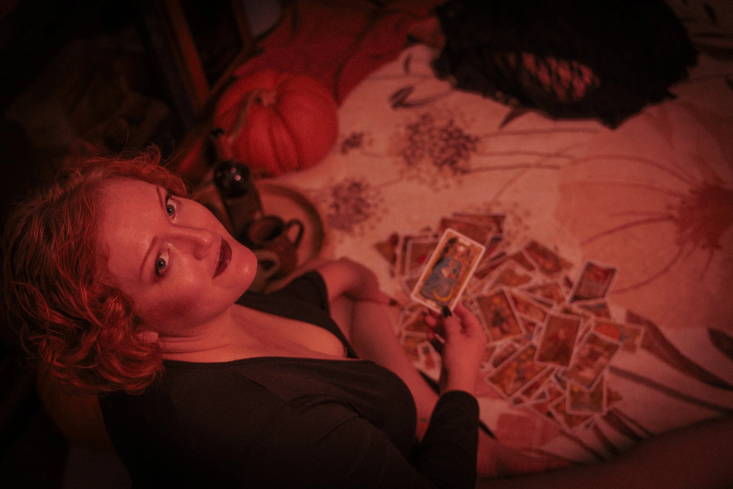 A woman with red curly hair and black top sitting on a floral bedspread, looking up at the camera, with tarot cards and Halloween decorations nearby.