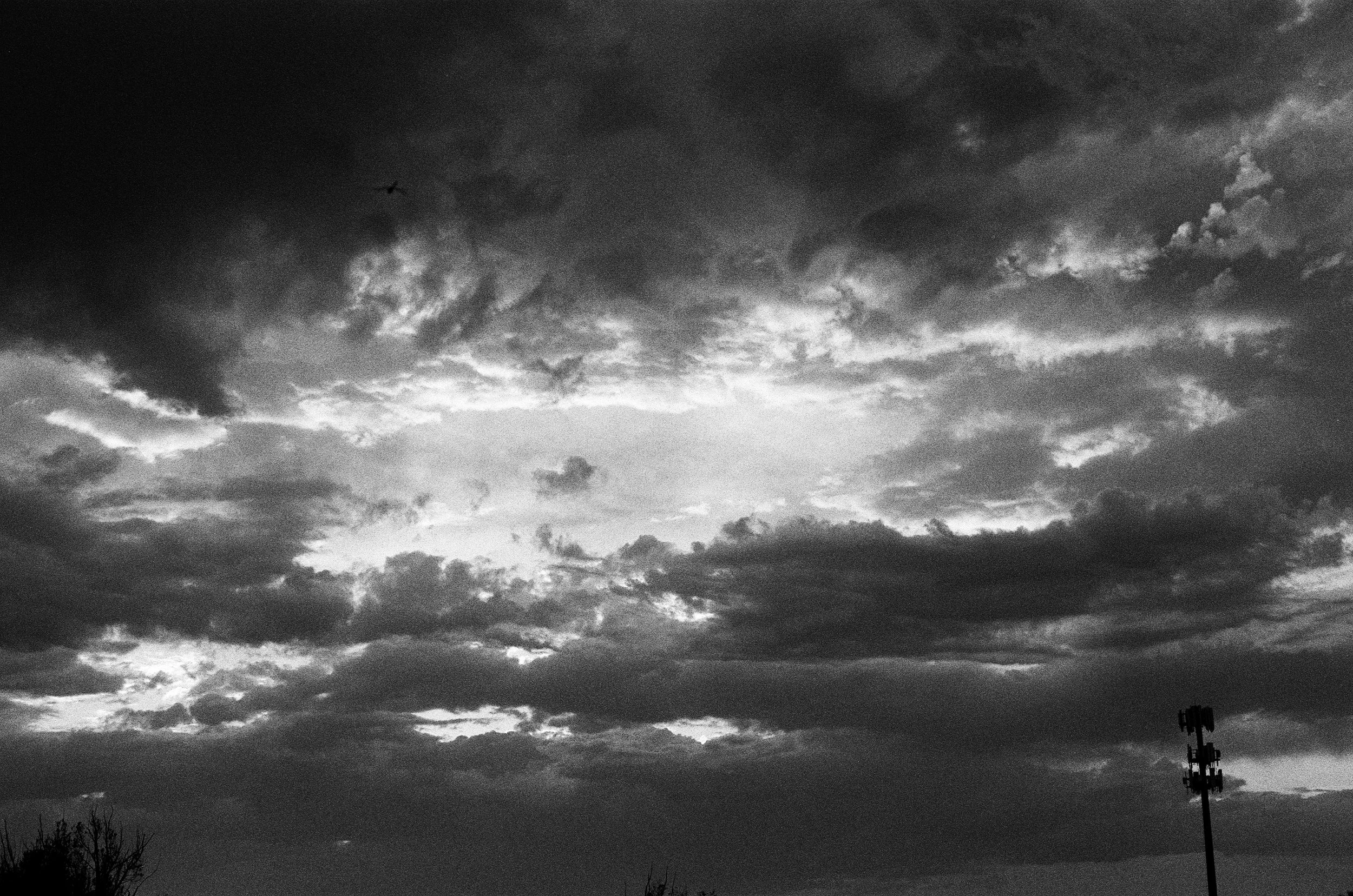 Black and white photo of a cloudy sky with a small airplane flying and a tall pole with lights in the foreground.