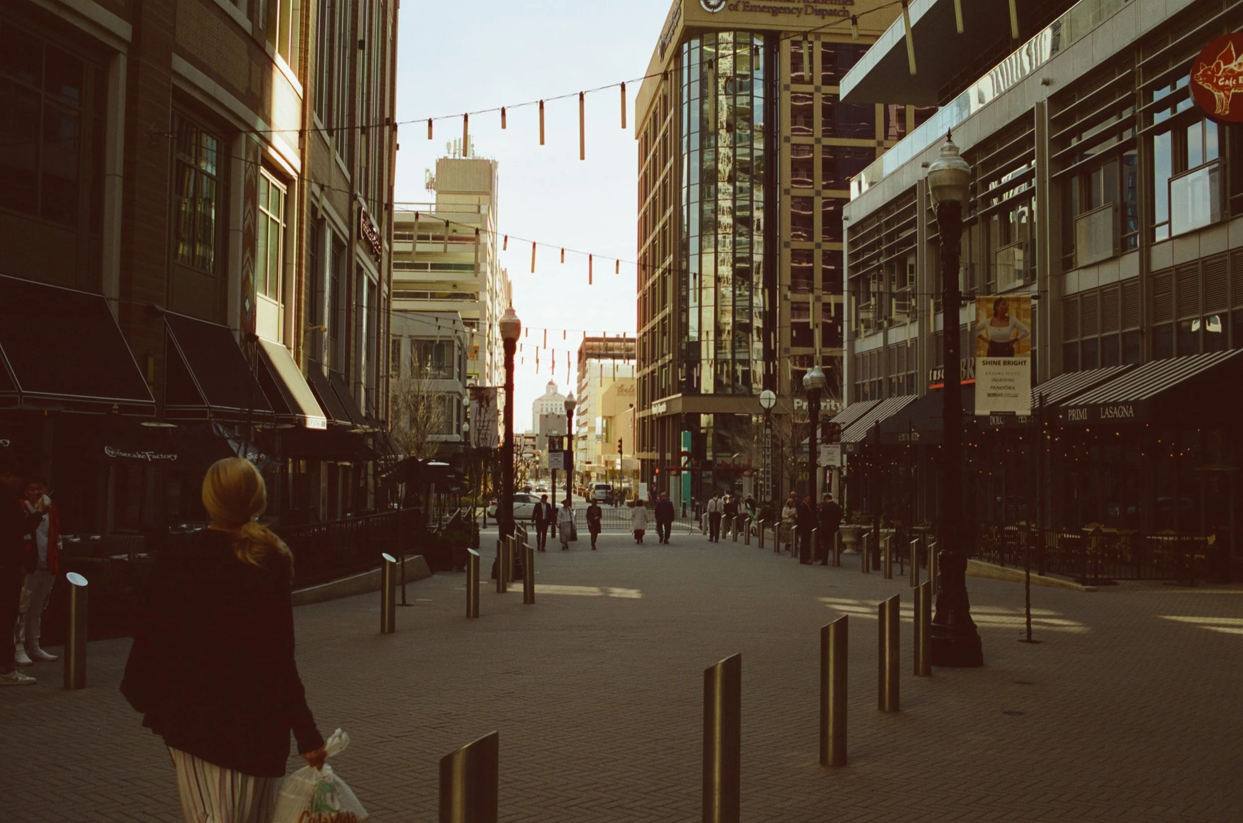 A busy city street with tall glass buildings, pedestrians walking, and string lights hanging across the street.