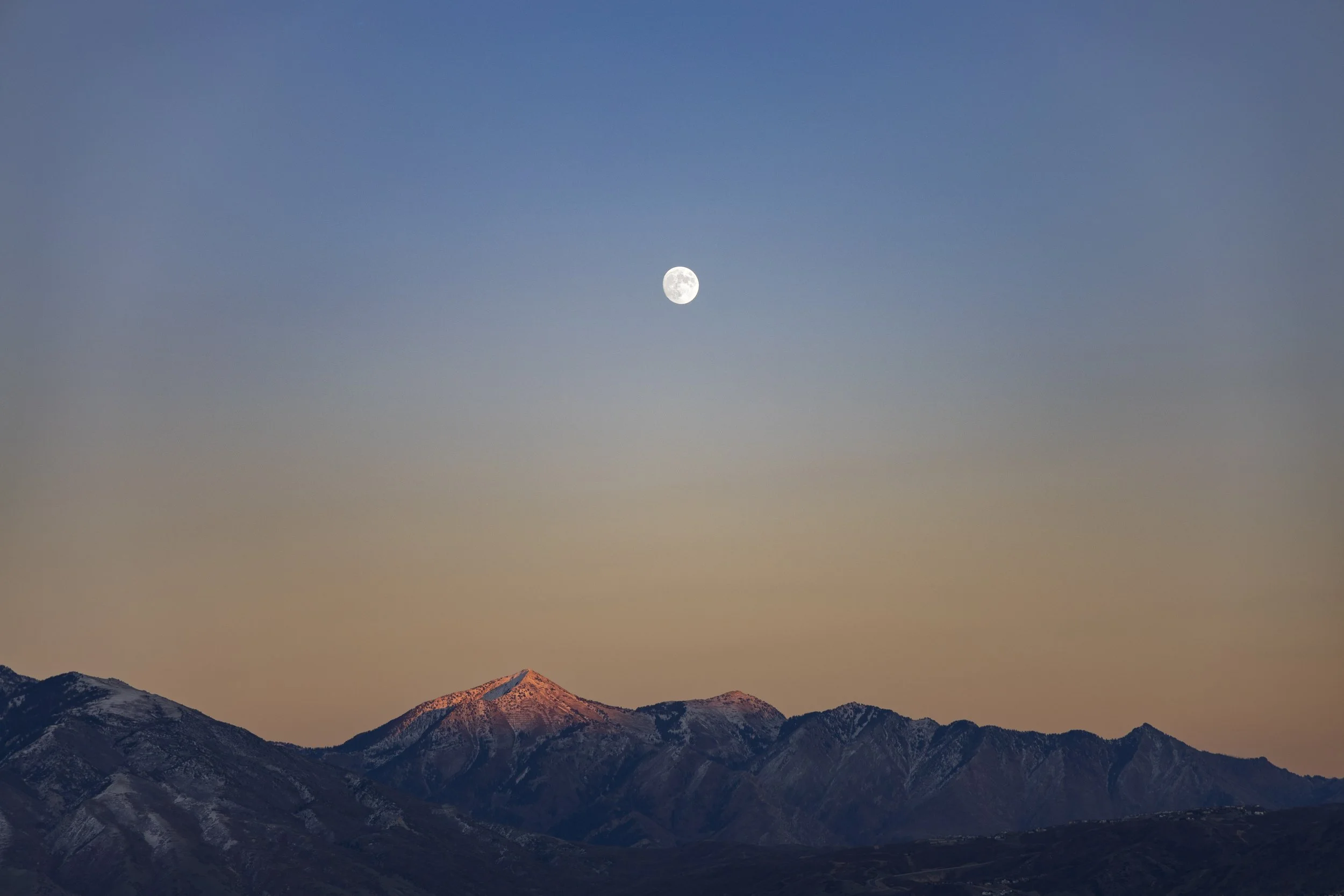 Mountains with snow-capped peaks at sunrise, with the moon visible in a clear blue sky.