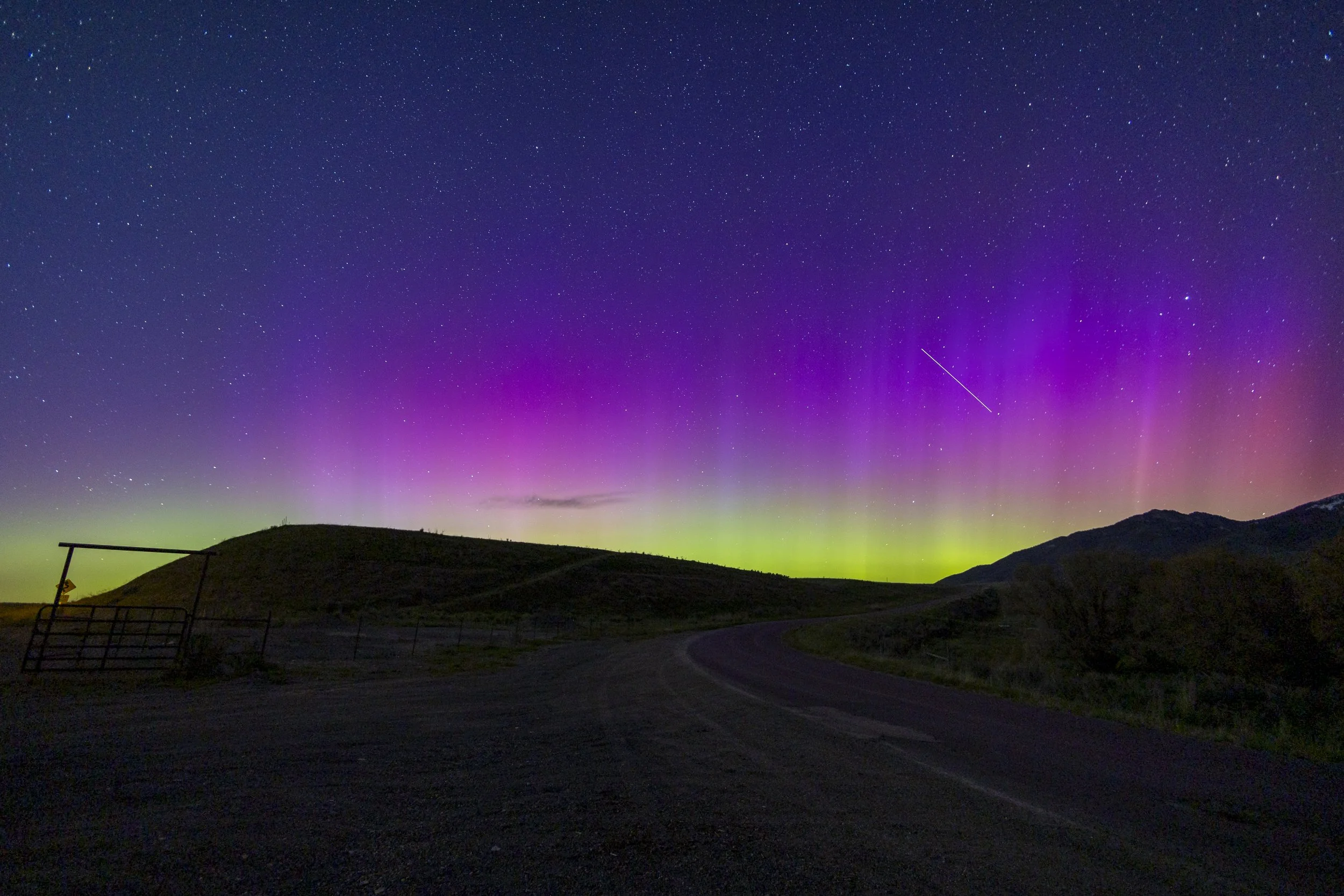Night sky illuminated with colorful aurora borealis and stars, with a silhouette of rolling hills and a curved road in the foreground.