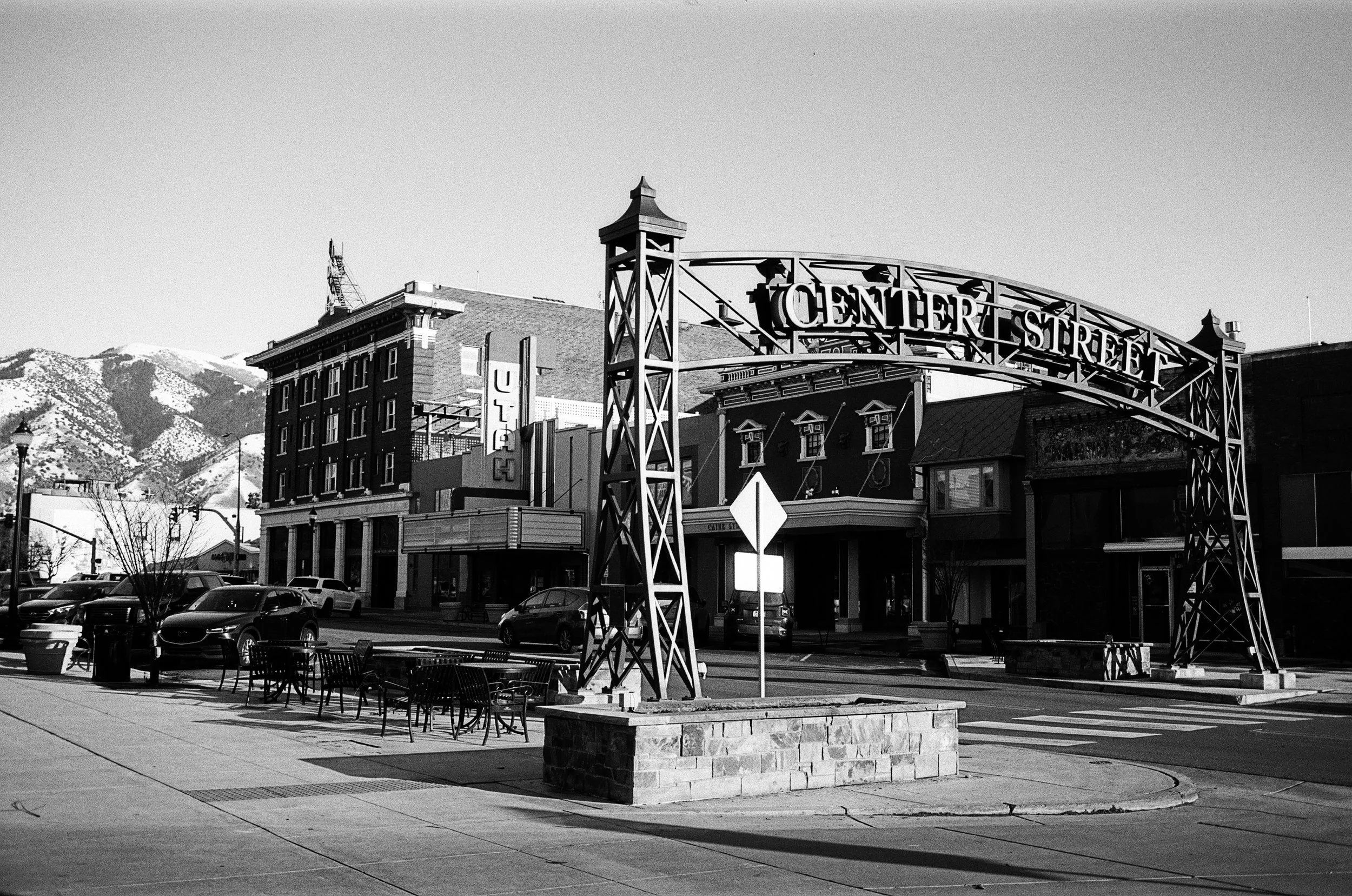 Black and white photo of Center Street sign over a street with parked cars, buildings, and snow-capped mountains in the background.