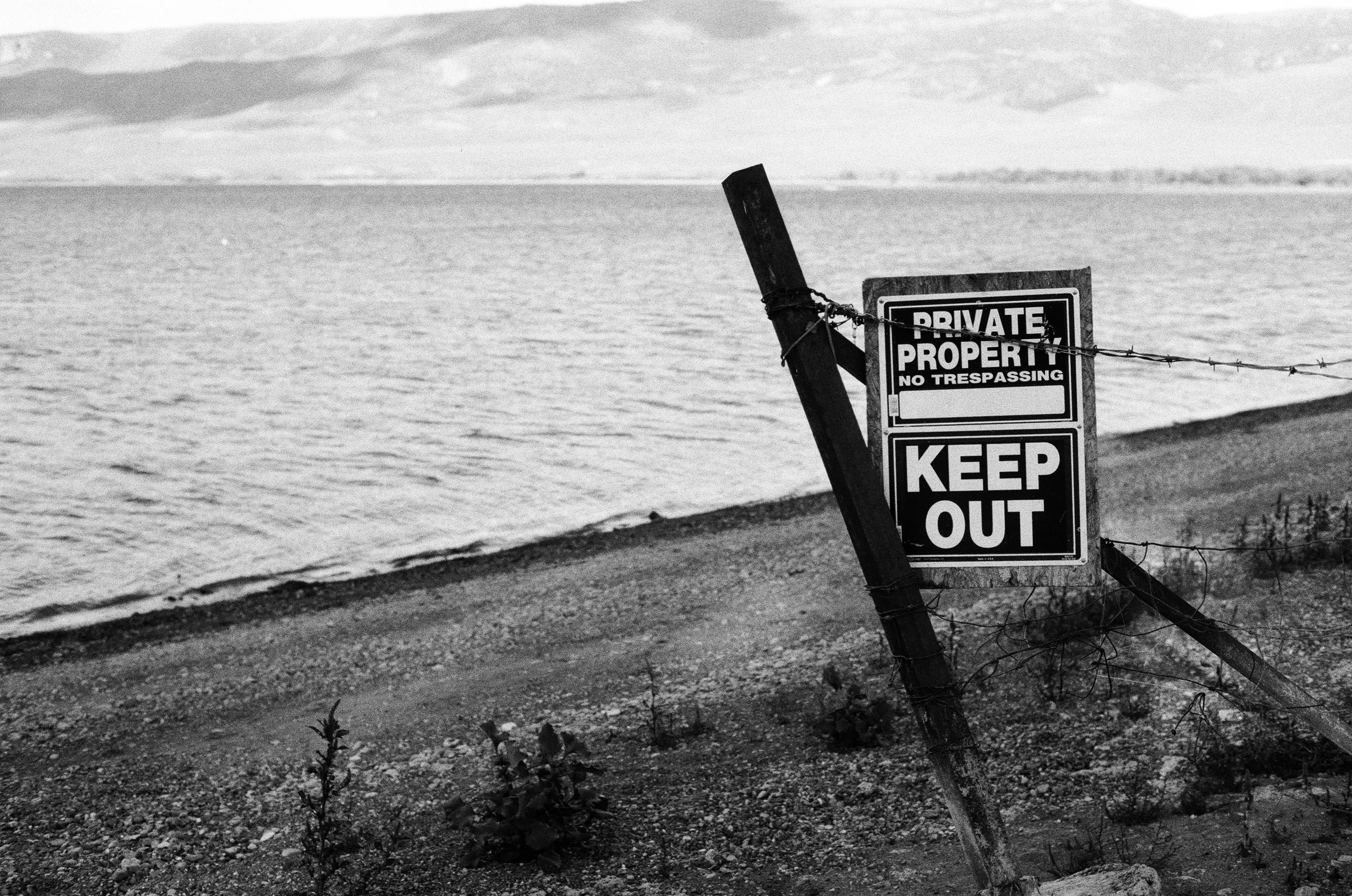 A black and white photograph of a shoreline with a leaning wooden post holding a sign that reads 'PRIVATE PROPERTY NO TRESPASSING KEEP OUT'. The sign is attached to a barbed wire fence, and there is a body of water with calm waves in the background.
