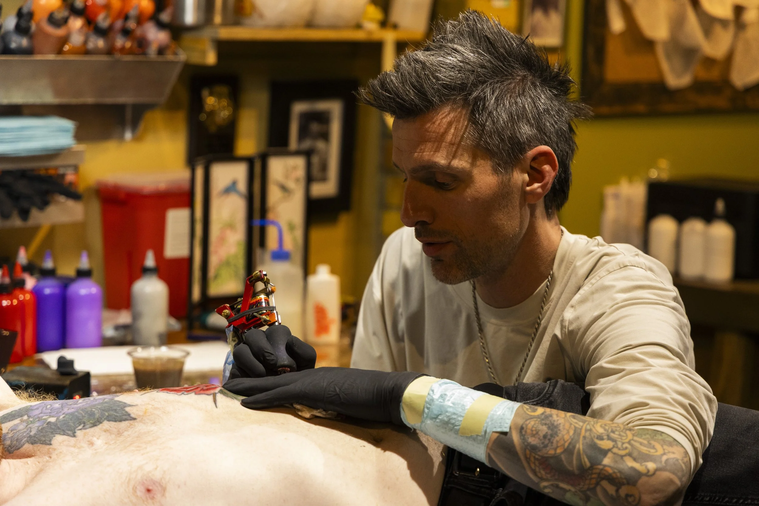 A man with tattoos receiving a tattoo from an artist in a tattoo shop.