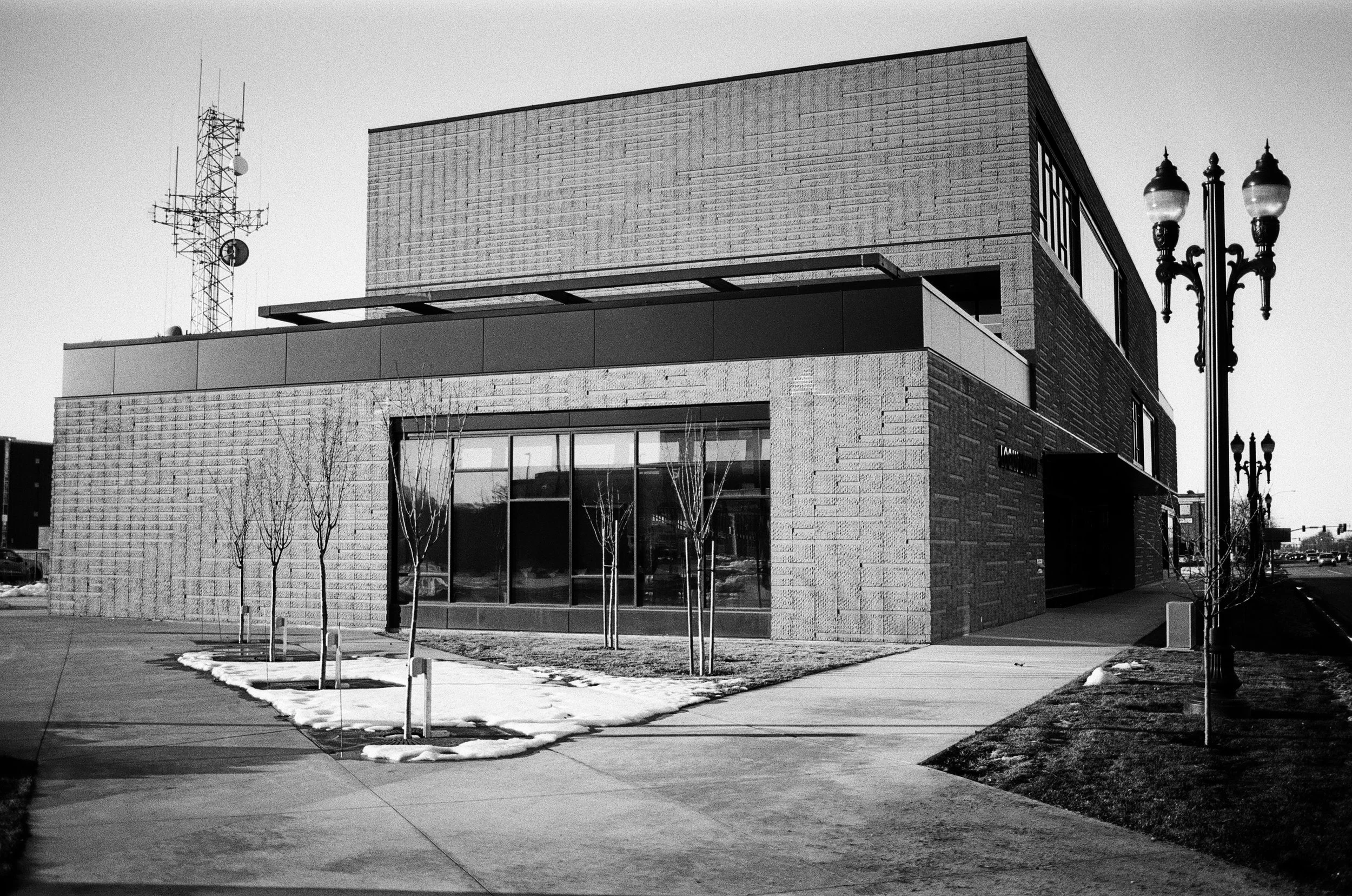 Modern brick building with large glass windows, lamp posts, and leafless trees in front, with patches of snow on the ground, on a partly cloudy day.