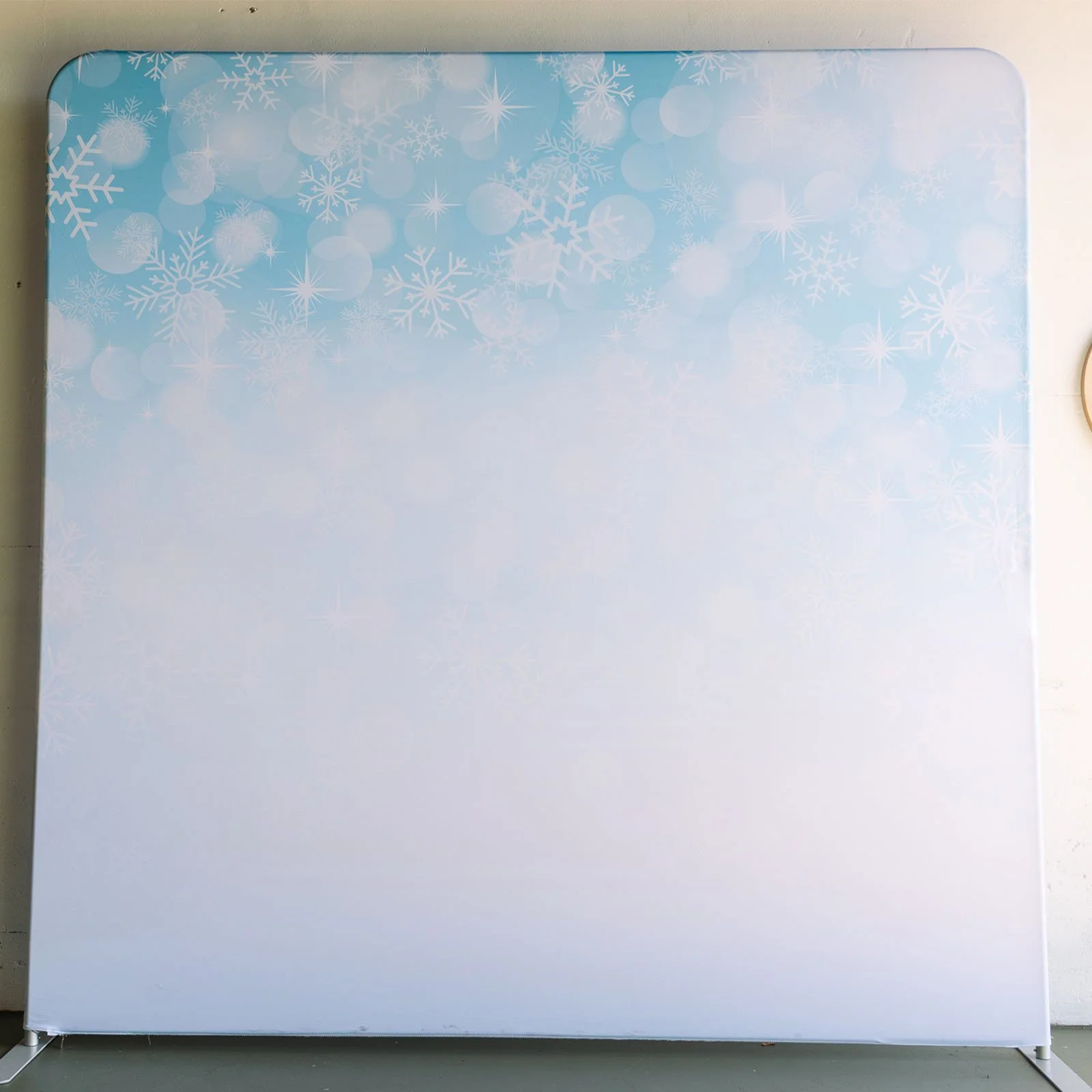 A white panel or backdrop with a light blue top border decorated with white snowflakes and bokeh light effects, used for photo shoots or displays.