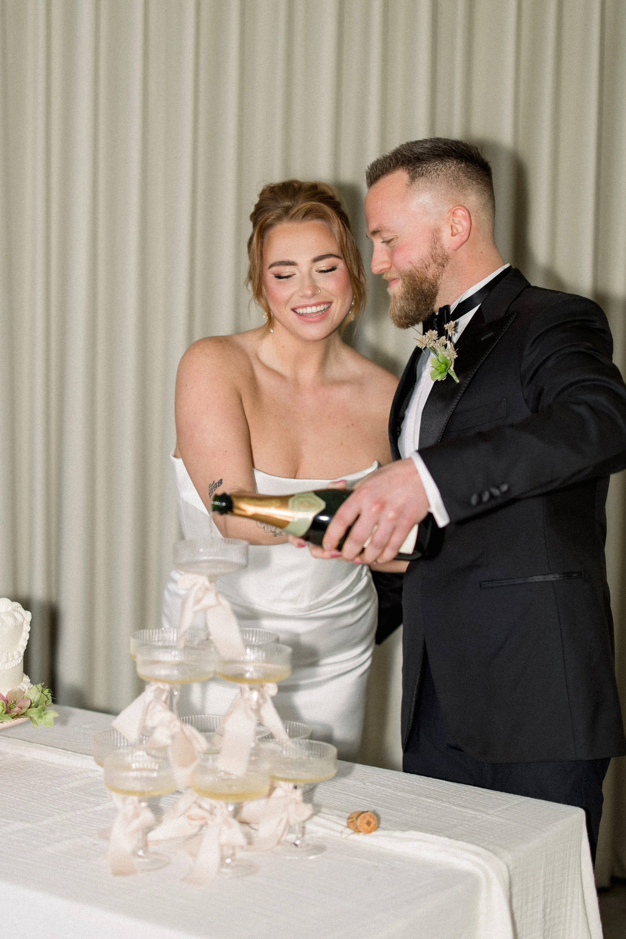 Bride and groom in wedding attire opening a champagne bottle at their wedding reception.