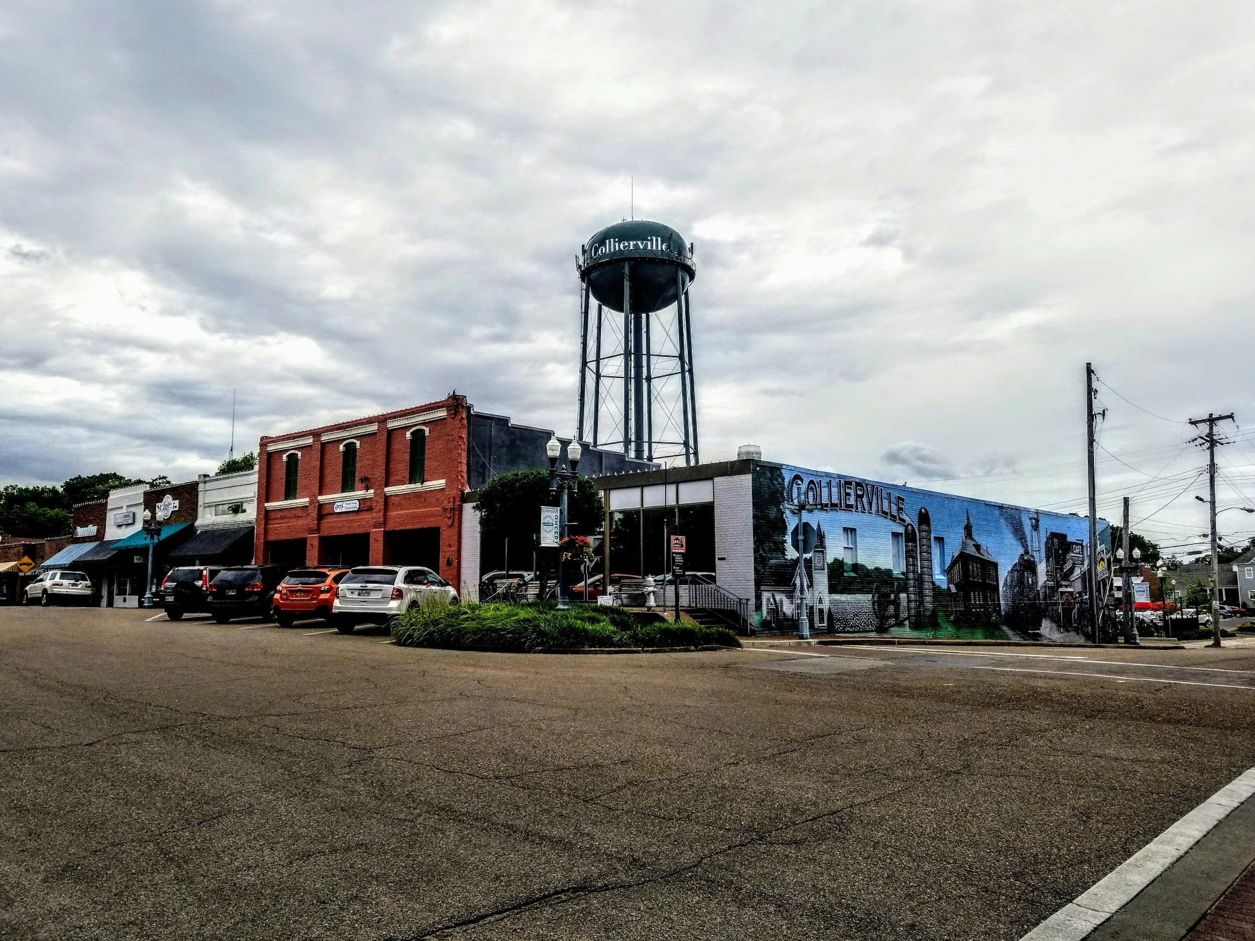 A small-town street scene with historic brick buildings, a water tower with the word 'Collierville,' parked cars, and power lines under a cloudy sky.