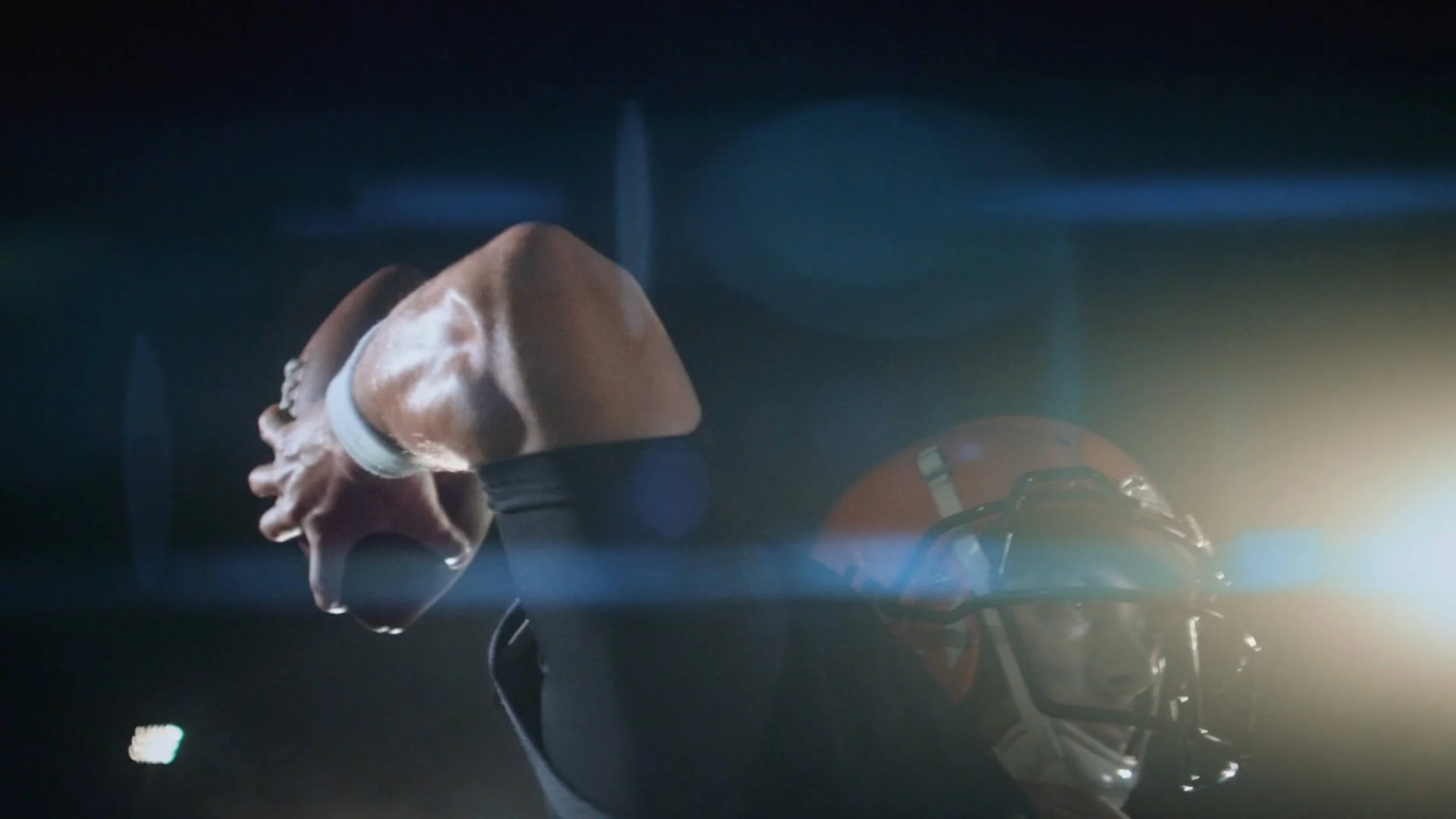 A football player in a dark setting with a helmet, illuminated by bright light from the side, and holding a football in both hands.