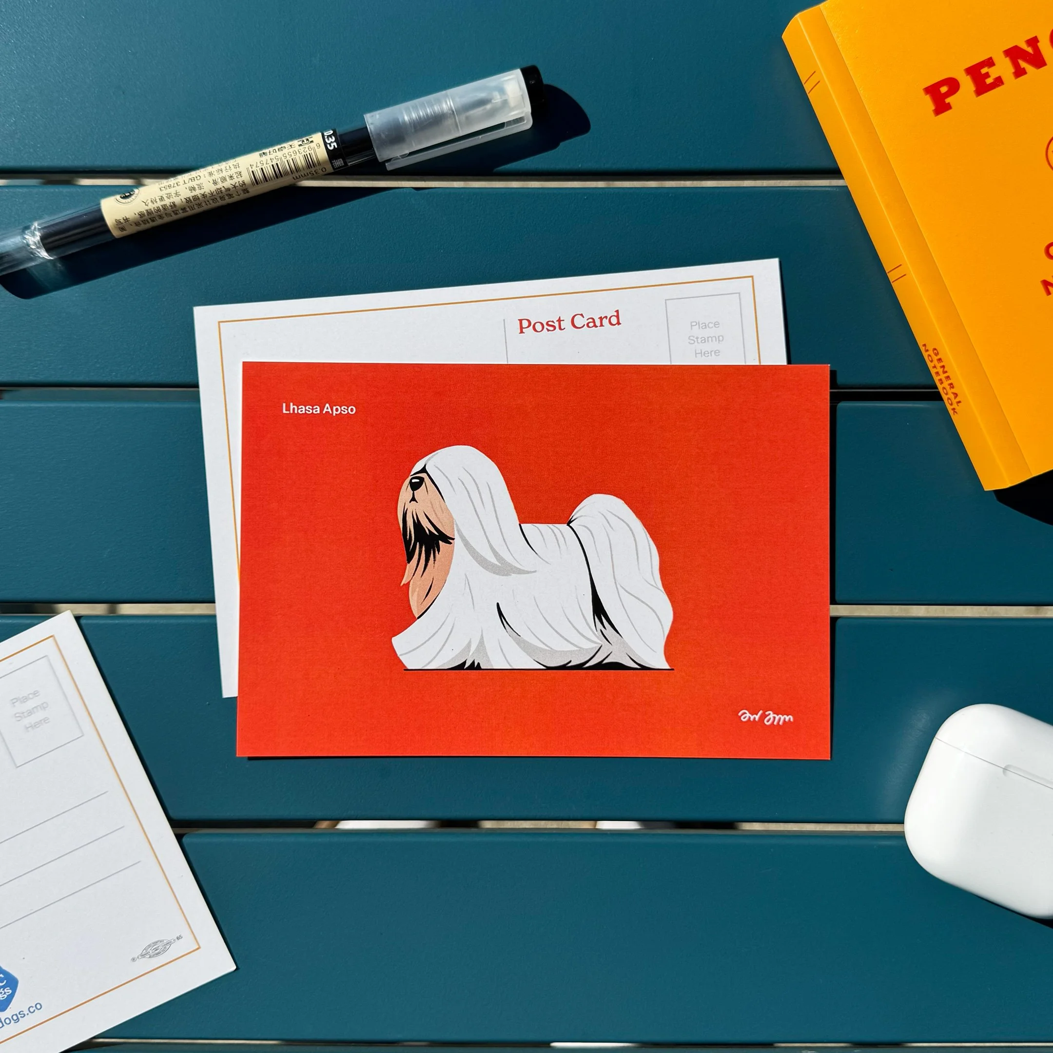 A red postcard featuring an illustration of a Lhasa Apsoand the artist's signature in the bottom right corner. Surrounding items include a white envelope, a black and beige pen, a yellow book, and a pair of AirPods on a blue table