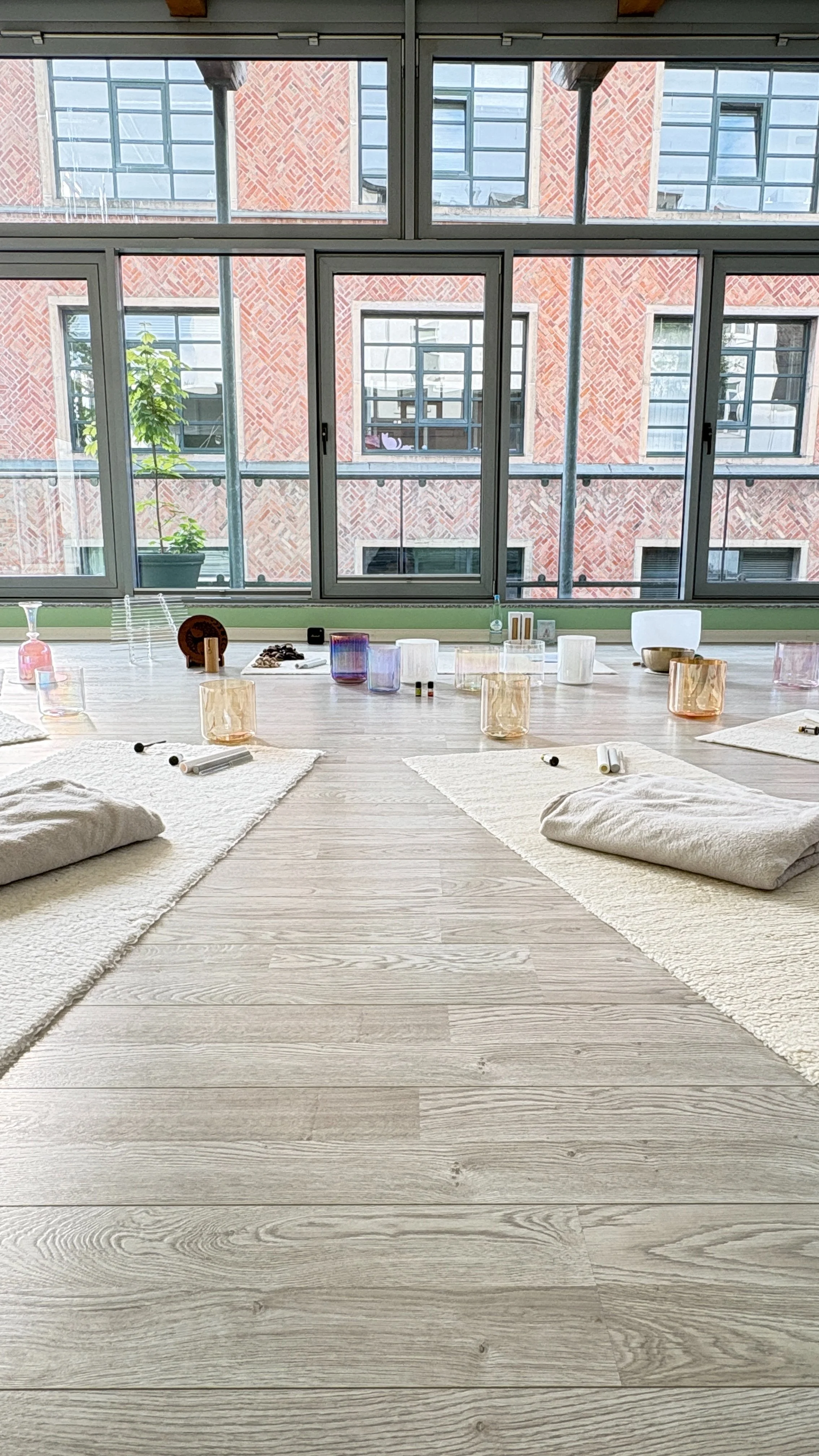 Vorbereiteter Raum für ein Soundbath mit Yogamatten, Kissen und mehreren Kristallklangschalen vor großen Fenstern.