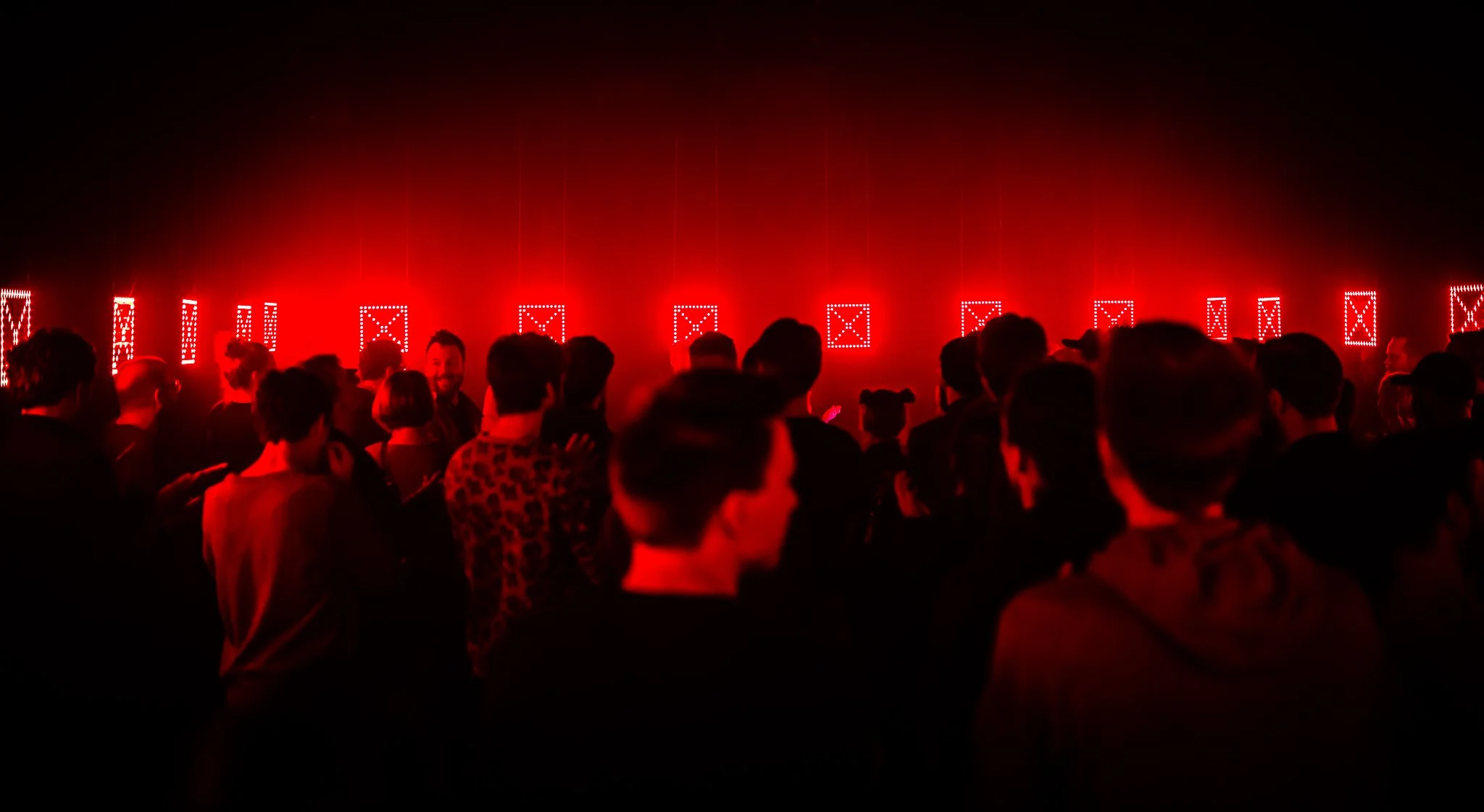 Publikum sett fra ryggen i rødt lys på konsert, med lysinstallasjon av firkantede LED-symboler hengende fra taket i bakgrunnen.